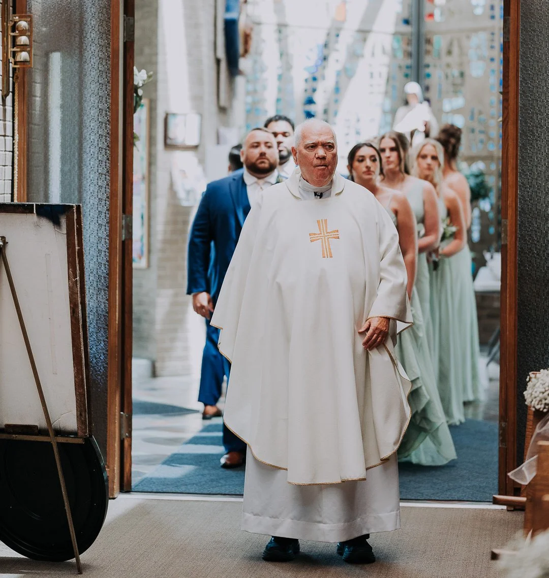 A priest leading a wedding procession with the bride and groom and bridesmaids and groomsmen inside a church.