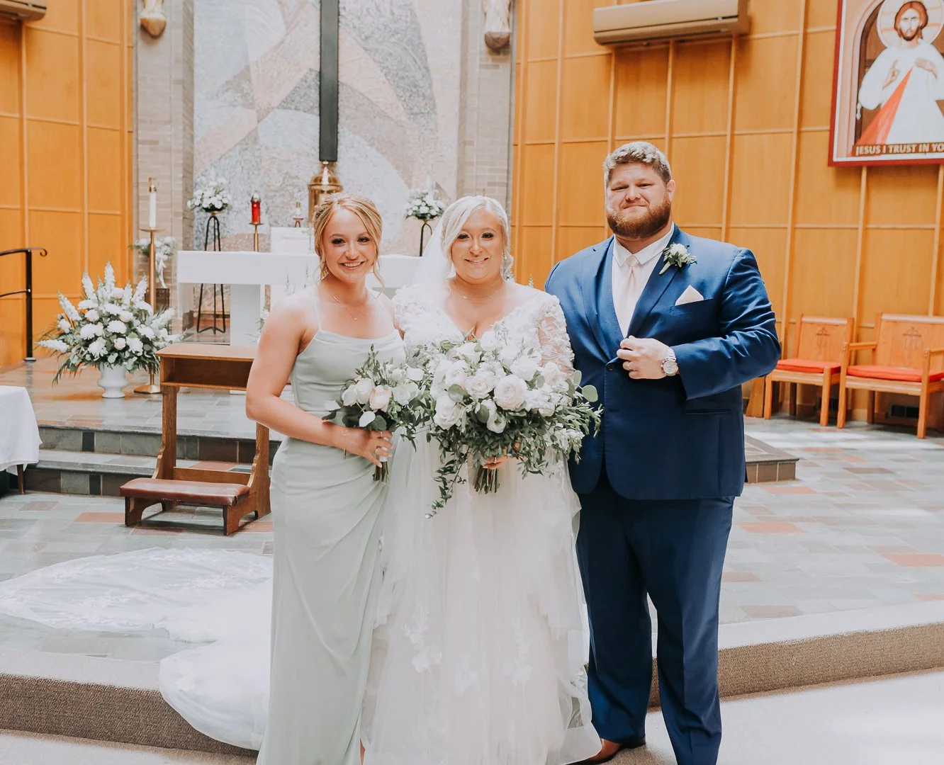 A bride and groom with a bridesmaid standing inside a church. The bridesmaid is holding a bouquet, and the bride has a bouquet of white roses and greenery. The church has wooden walls, chairs, and religious artwork.