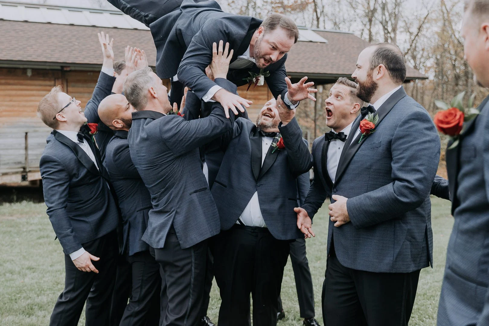A group of men in suits and tuxedos celebrating outdoors, lifting a man in a suit above their heads during a wedding or similar event.