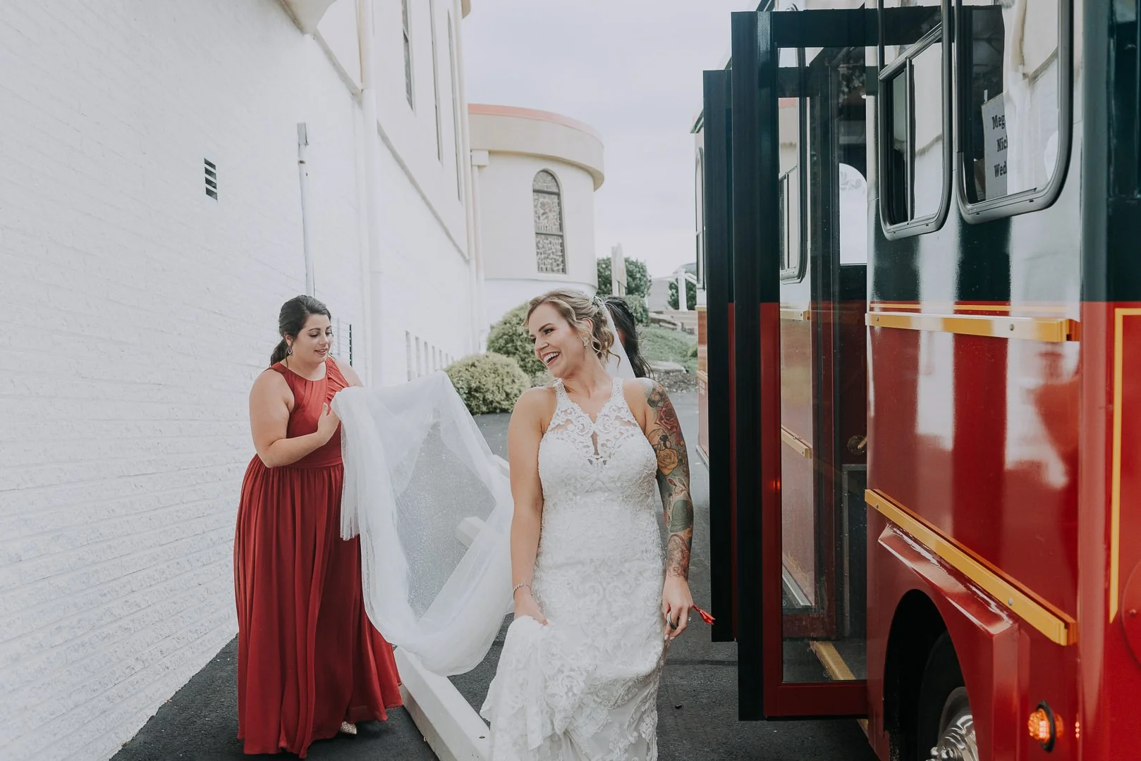 Bridesmaid in red dress helping bride in white wedding gown with veil near a red food truck outdoors.