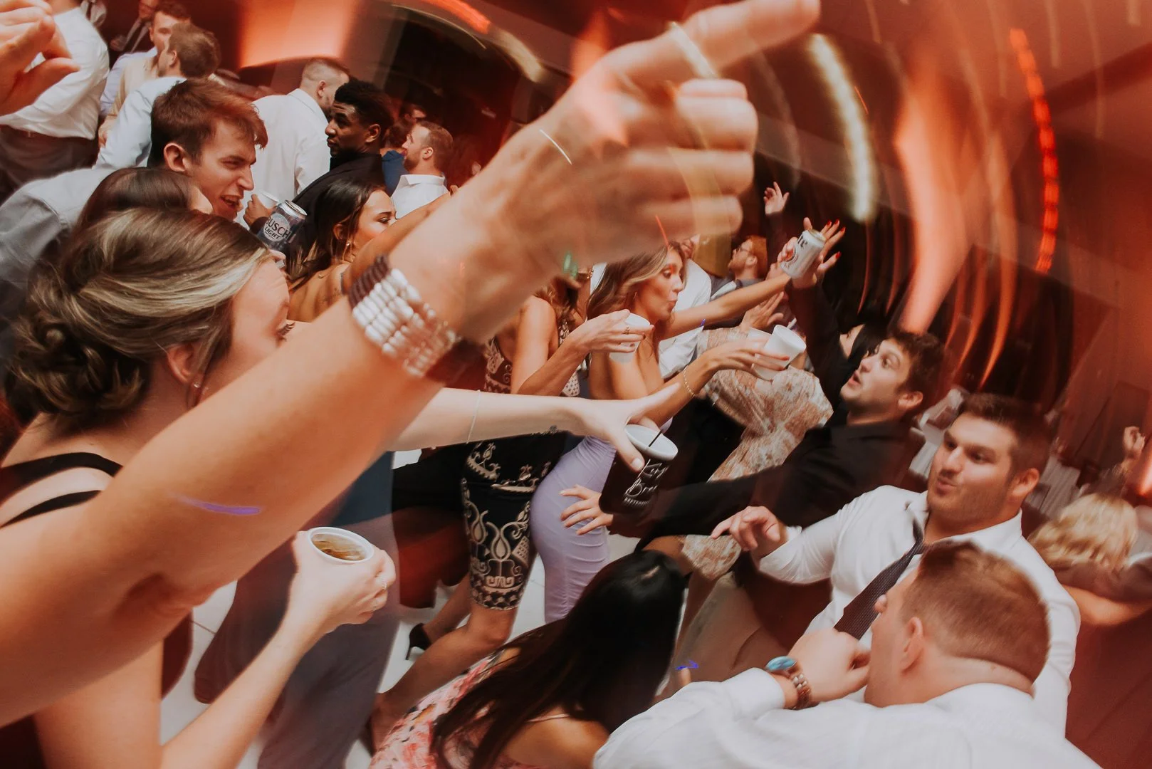 A lively party scene with people dancing and socializing, holding drinks, with a woman in the foreground raising her hand, wearing bracelets, in a red-lit room.