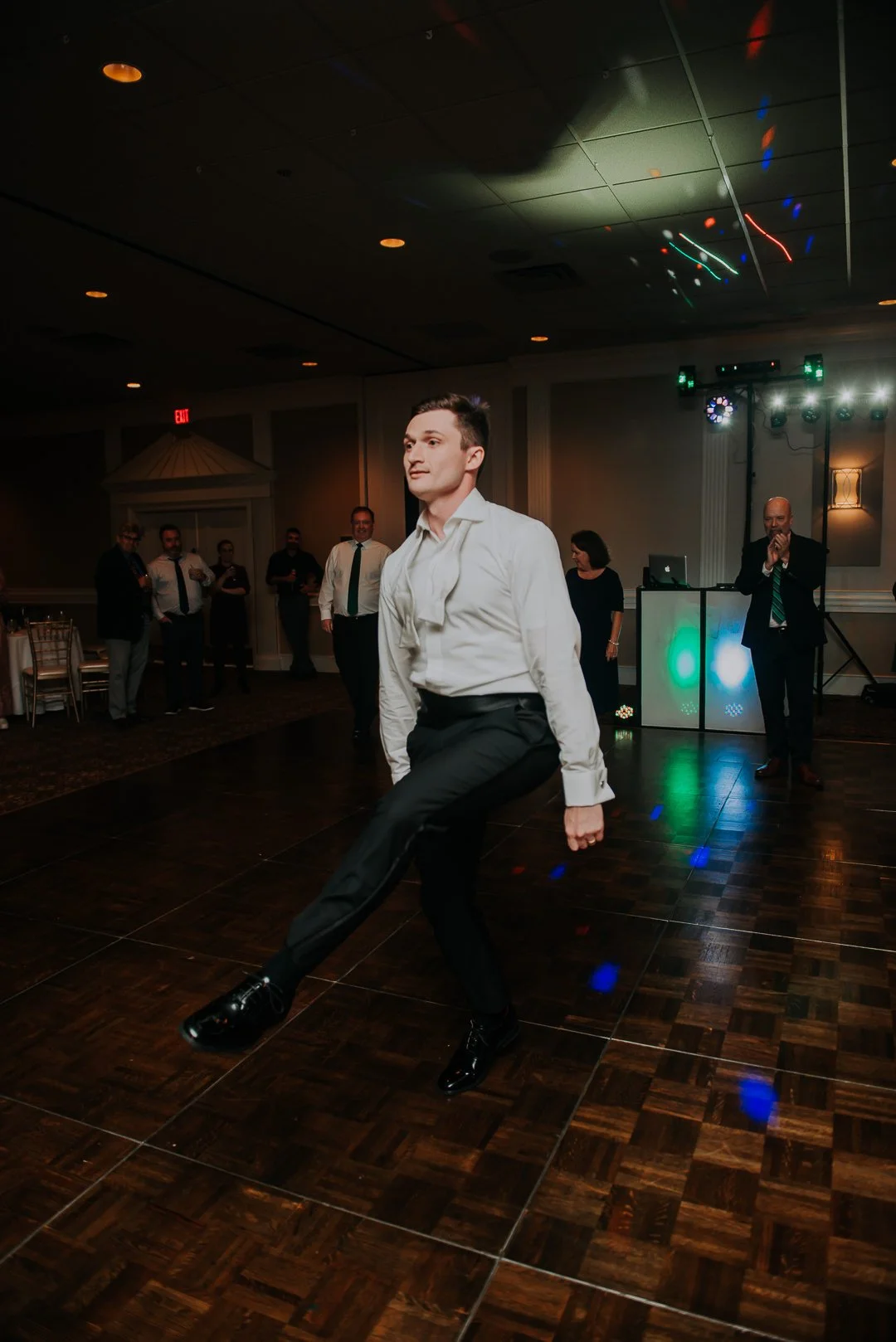 A man in a white shirt and black pants dancing at a formal event with a group of people in the background. The dance floor is wooden, and there are colorful lights and a DJ booth in the background.