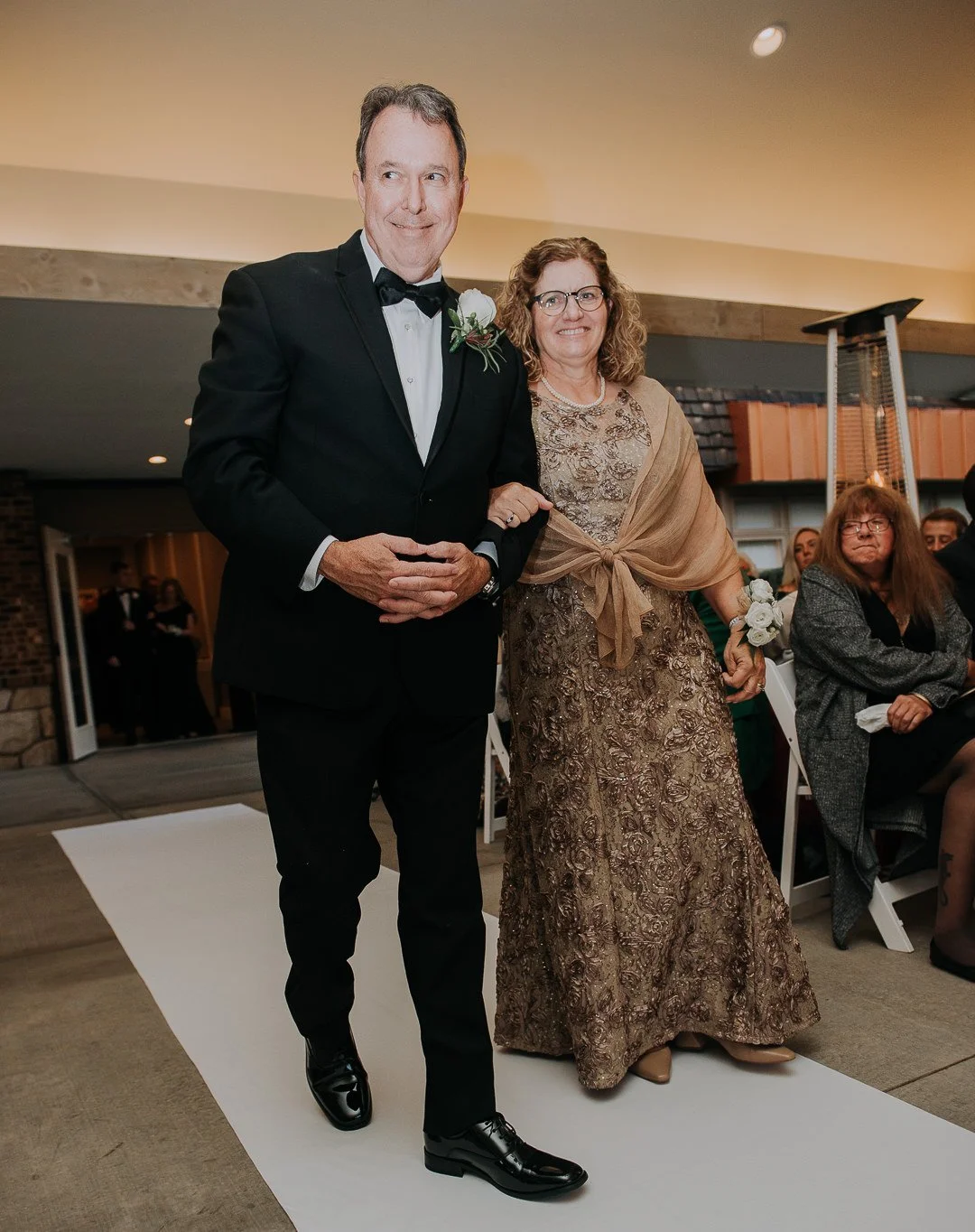 A groom in a black tuxedo walking down the aisle with a woman in a gold dress and beige shawl, holding hands at a wedding ceremony.