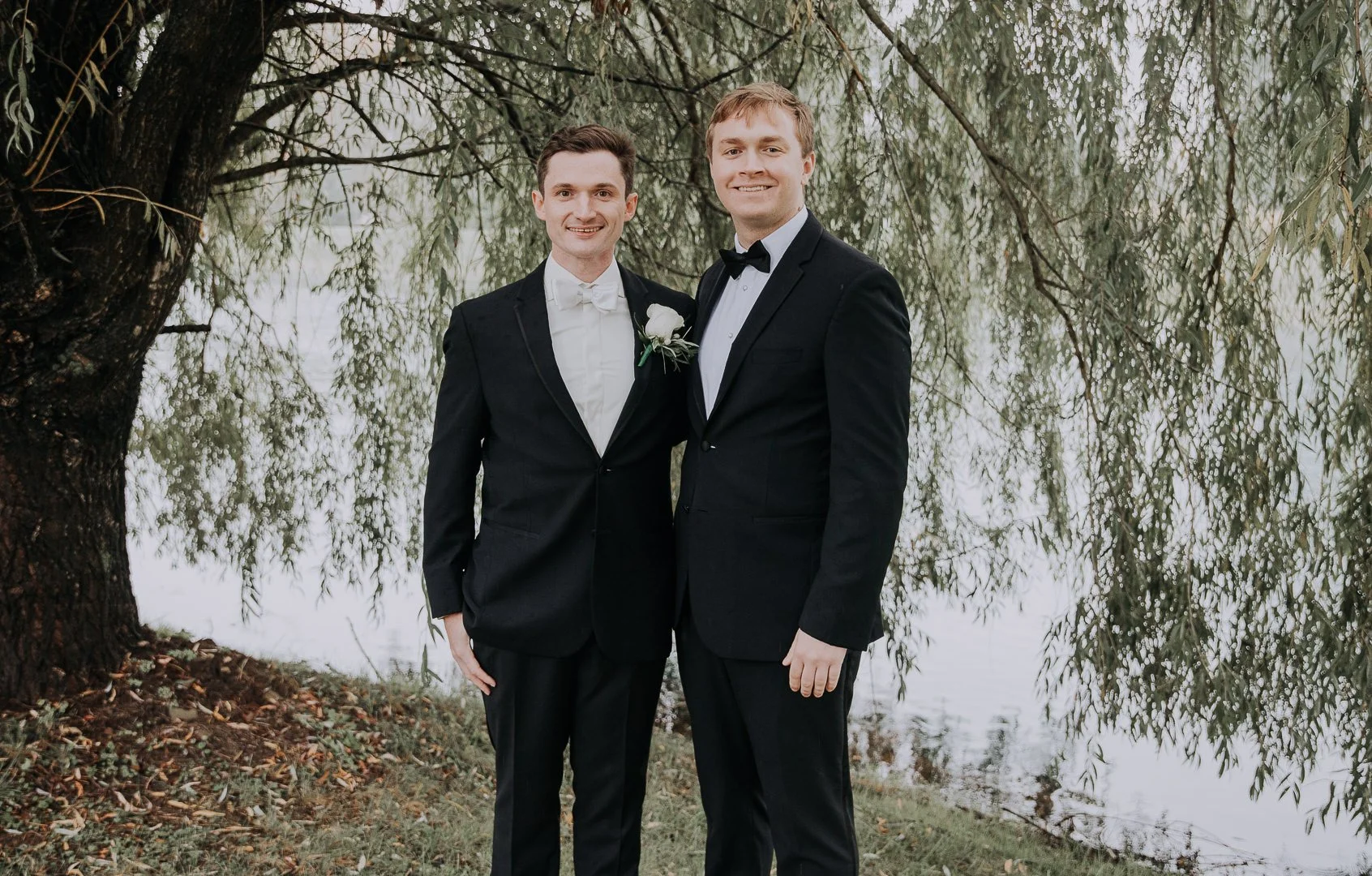 Two men in tuxedos standing outdoors near a large tree and a body of water, smiling at the camera.