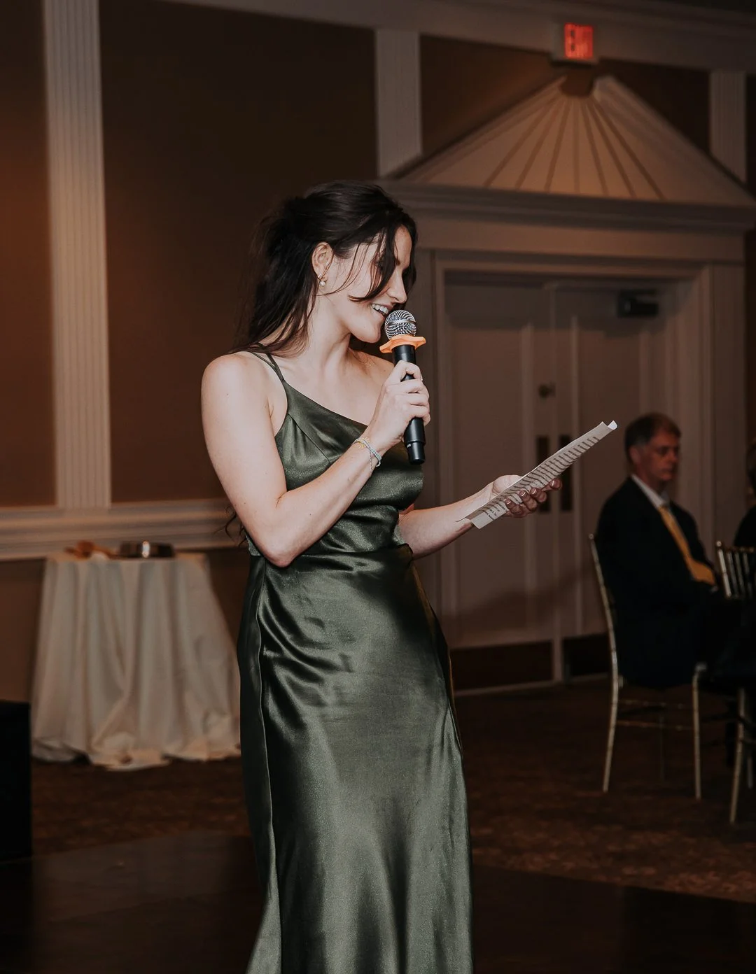 A woman in a green satin evening gown speaking into a microphone while reading from a paper during an indoor event.