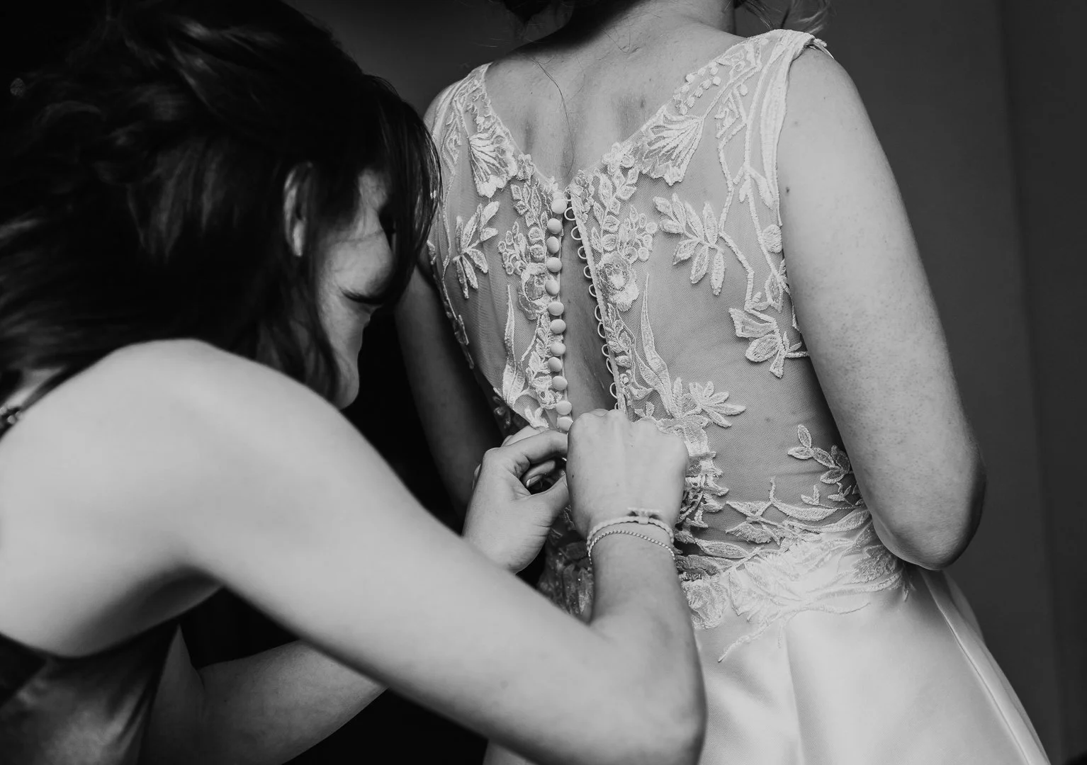 A woman helping another woman button up the back of her wedding dress with lace embroidery.