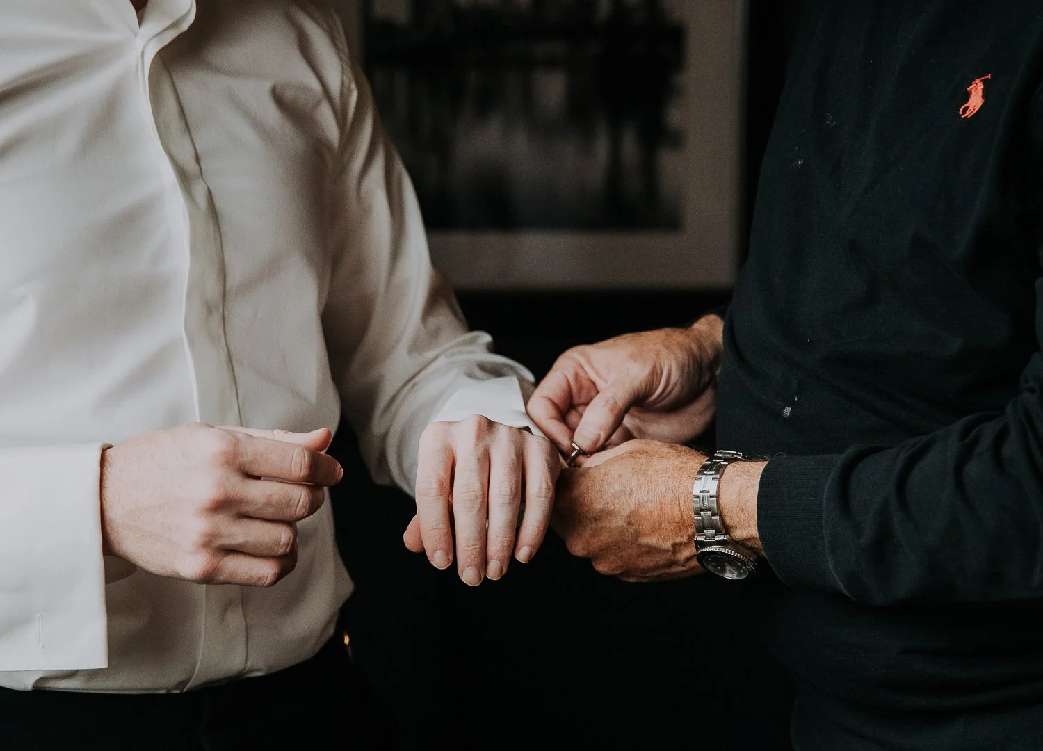 Two people are holding hands, with one person's hand being held by the other. The person on the right is wearing a black long sleeve shirt with a watch, and the person on the left is wearing a cream-colored long sleeve shirt.