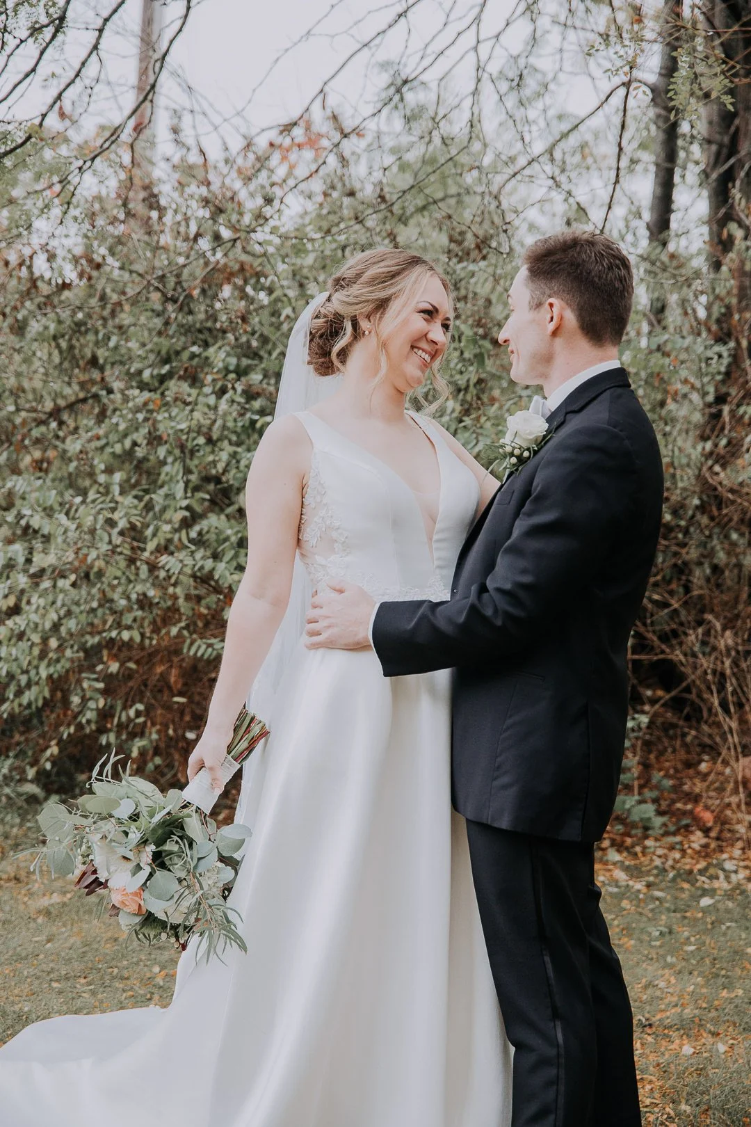 Bride and groom smiling and holding hands outdoors on their wedding day, with the bride holding a bouquet of greenery and flowers, and the groom in a tuxedo.