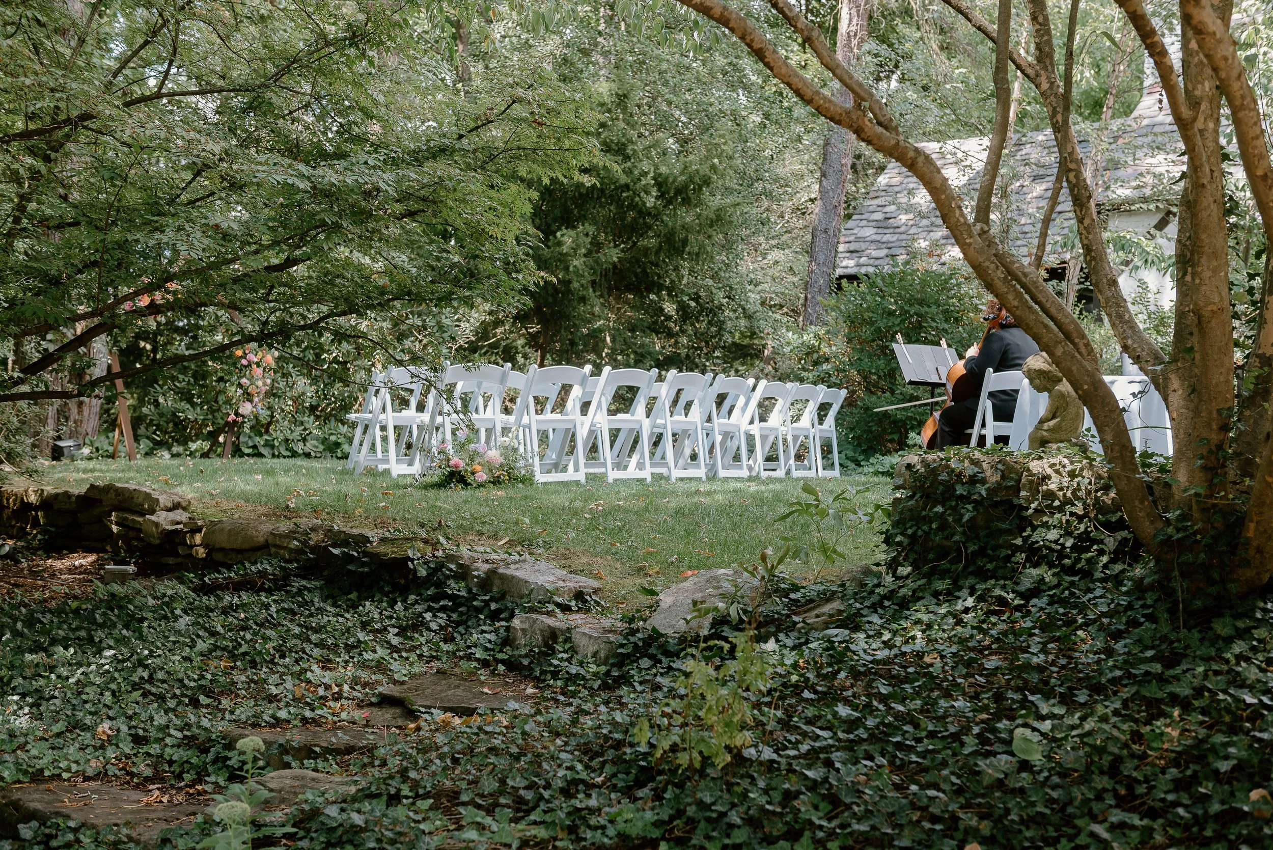 Outdoor wedding ceremony setup in a garden with white chairs arranged in rows, flowers on the ground, and a musician playing cello in the background among trees and shrubs.