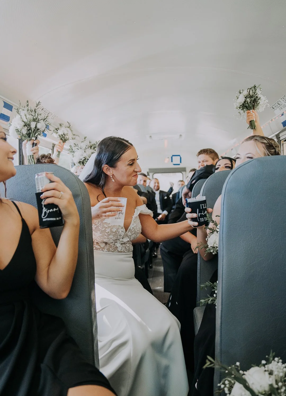 A wedding celebration inside a bus with a bride in a white dress reaching out to clink glasses with a guest. Guests are holding bouquets and drinks, and some are raising their hands in celebration.
