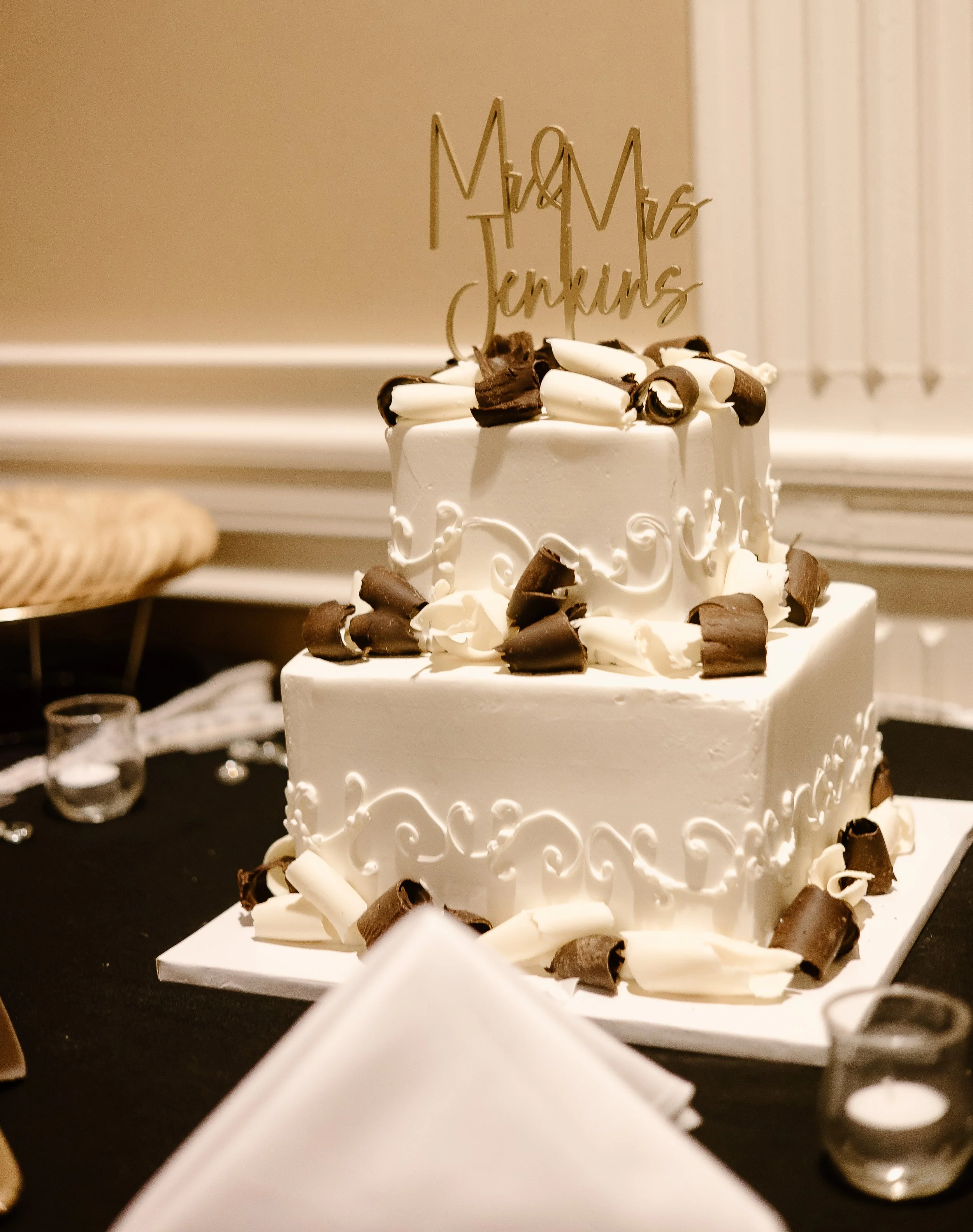 White two-tiered wedding cake decorated with chocolate curls and white swirls, topped with a gold 'Mr & Mrs' cake topper, on a black tablecloth, with candles and napkins nearby.