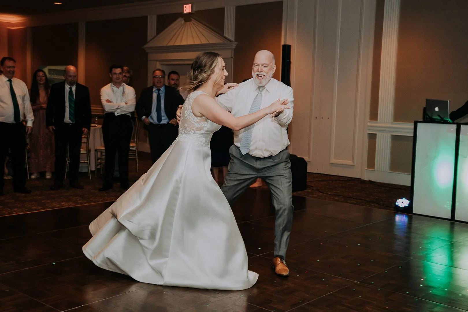A bride and an older man dancing together at a wedding reception, surrounded by guests watching and smiling.