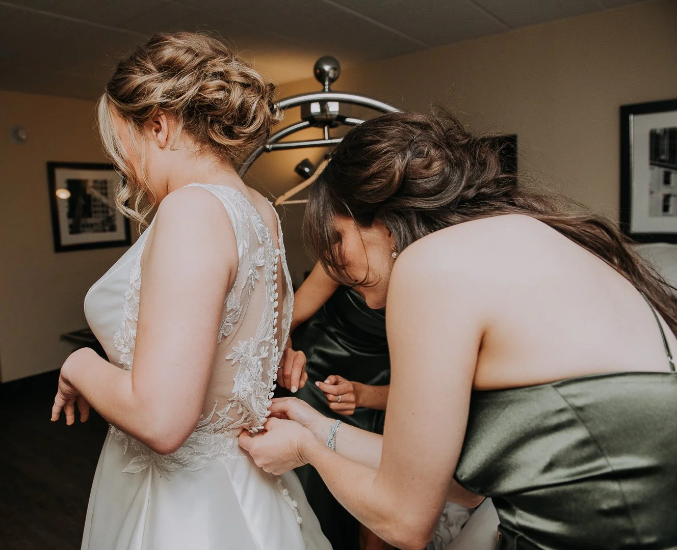 Women helping bride with wedding dress in a room with framed pictures on the wall.