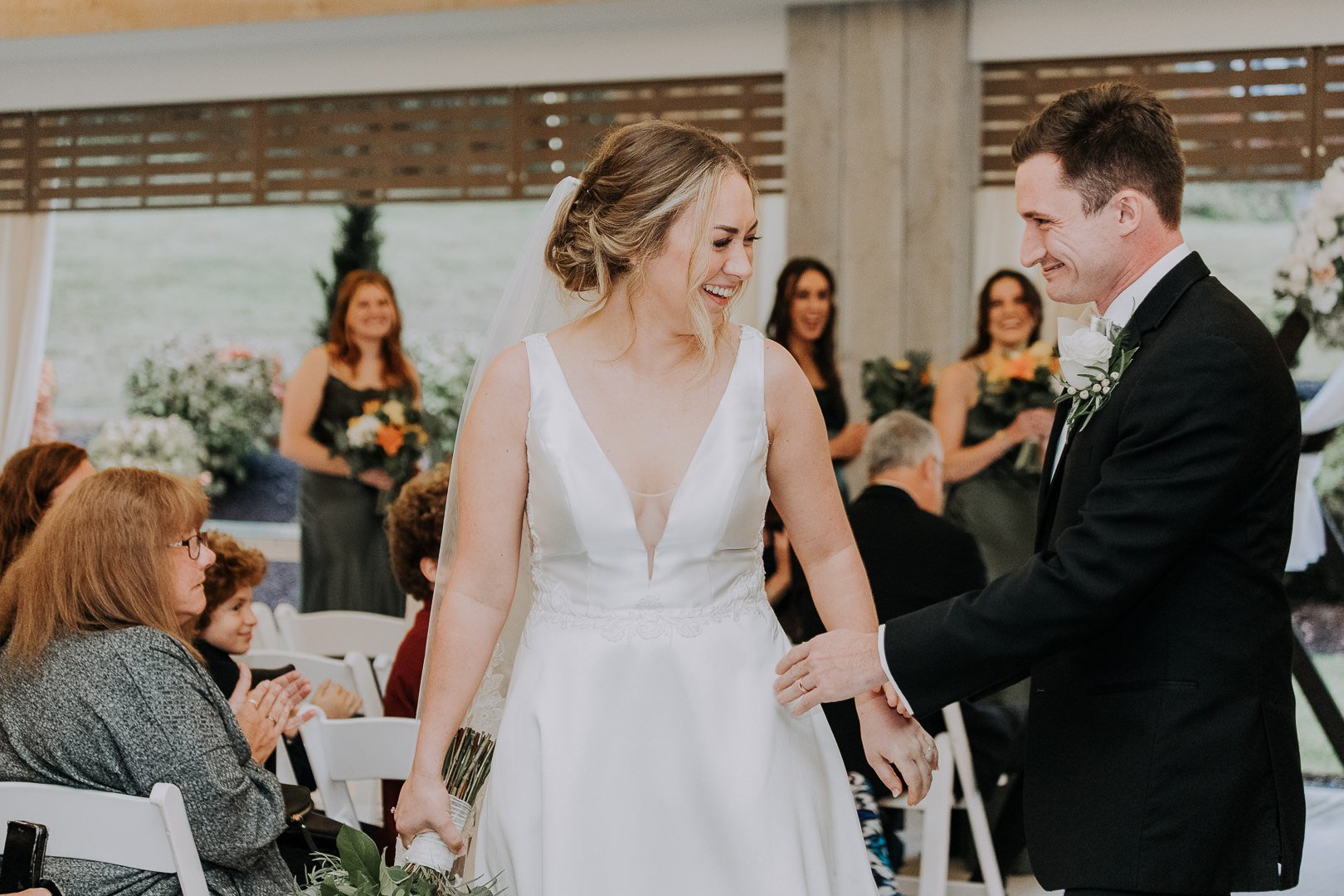 A bride and groom holding hands and smiling during their wedding ceremony, surrounded by seated guests and bridesmaids in a decorated venue.