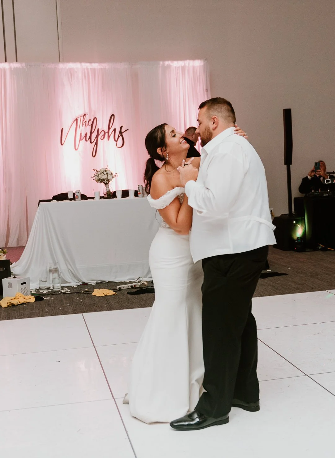 A bride and groom dancing at their wedding reception, with a pink backdrop and table in the background that says "The Tulips."