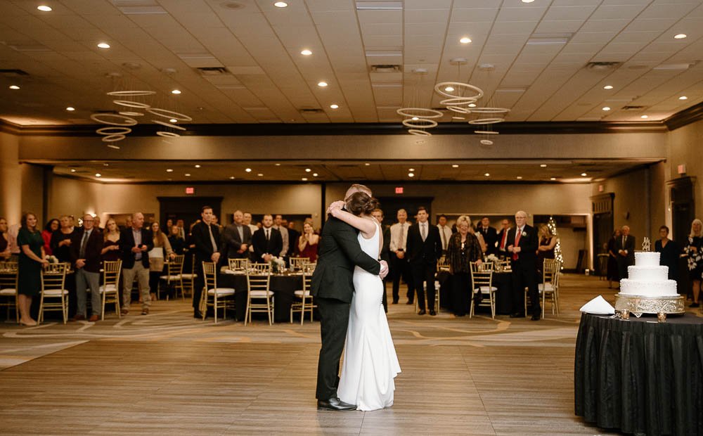 A couple shares a heartfelt dance at their wedding reception, surrounded by guests and elegant decor, with a wedding cake displayed nearby.