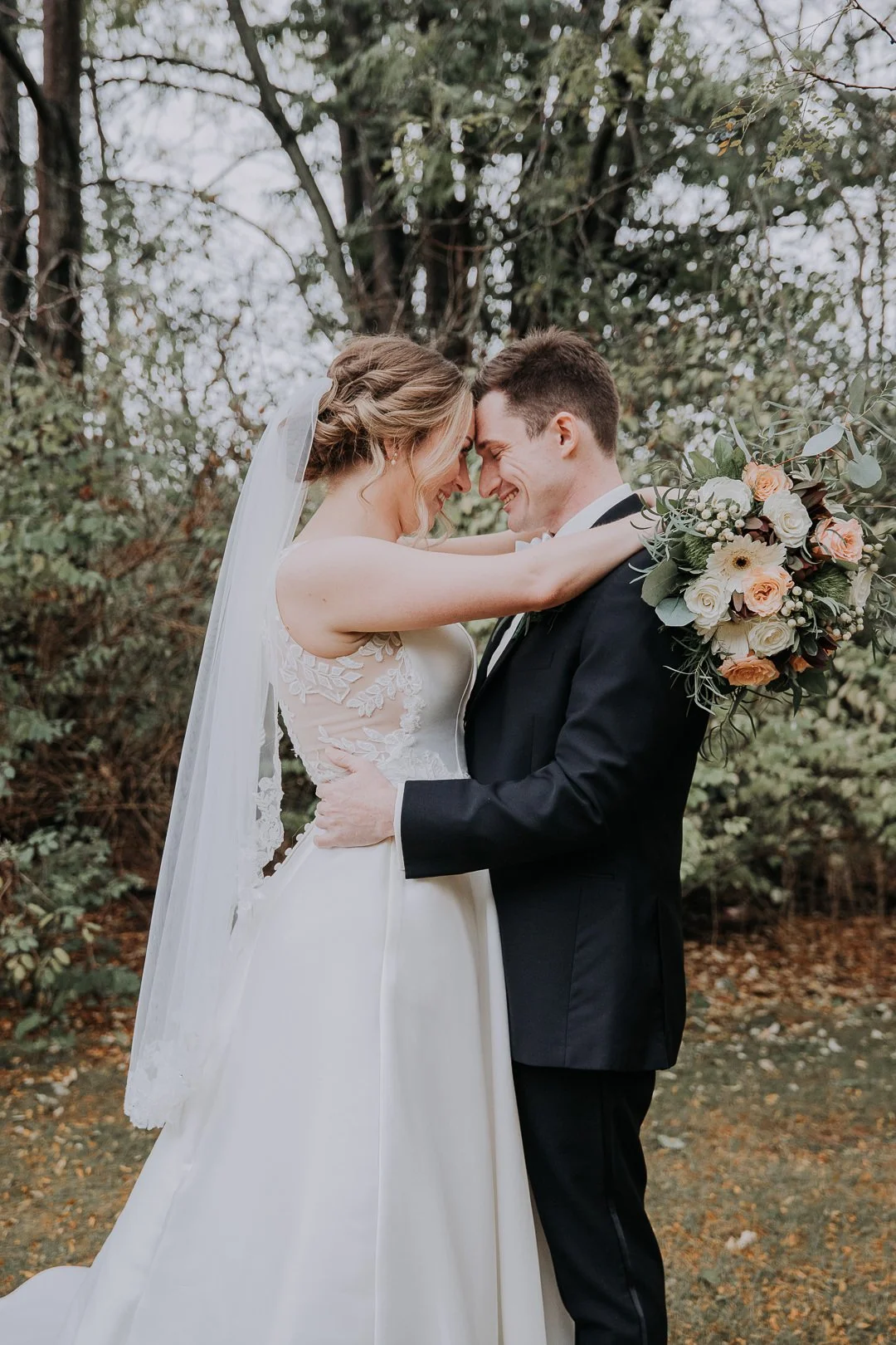 A bride and groom embrace outdoors during their wedding, smiling with their foreheads touching, with the bride holding a bouquet of flowers.