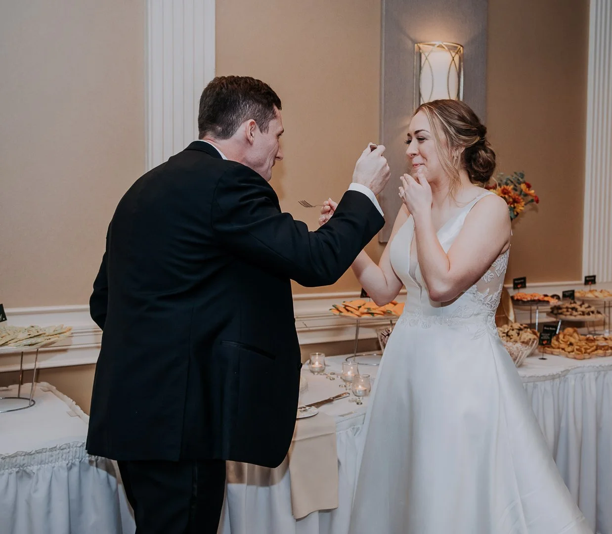 A groom feeding his bride at a wedding reception with a dessert table in the background.