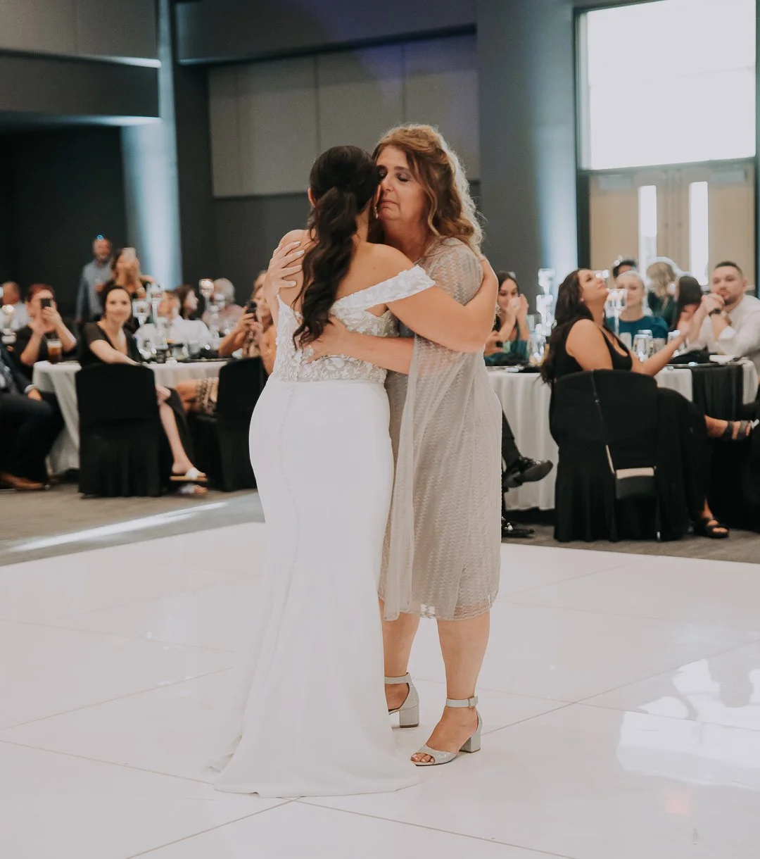 A bride and her mother share a dance at a wedding reception, with guests watching in the background.