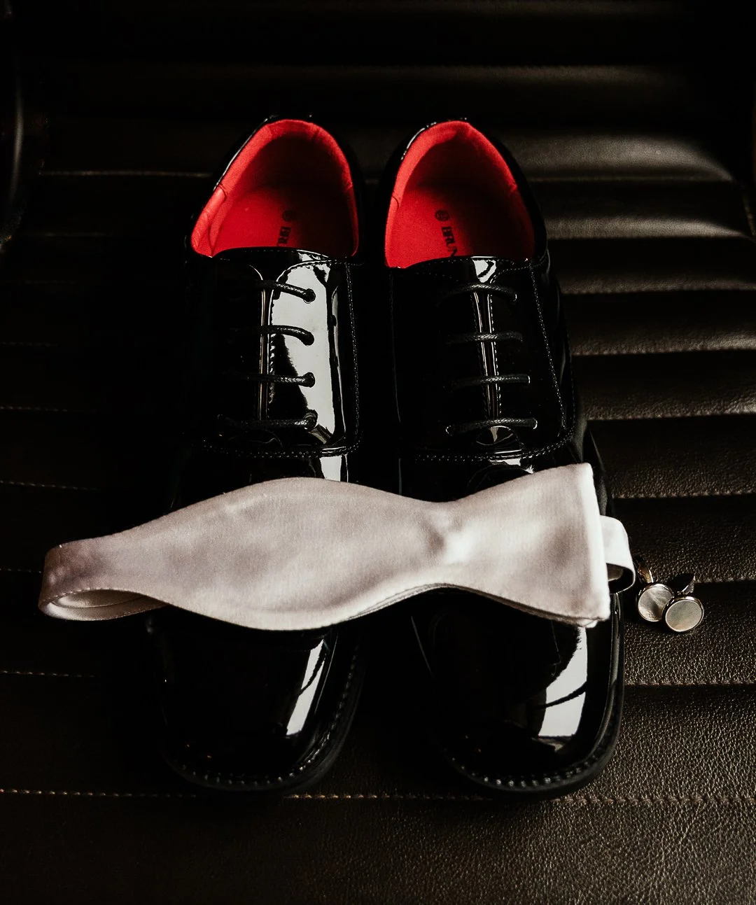 A pair of shiny black shoes with red insoles, a white cloth, and a pair of cufflinks on a black leather surface.