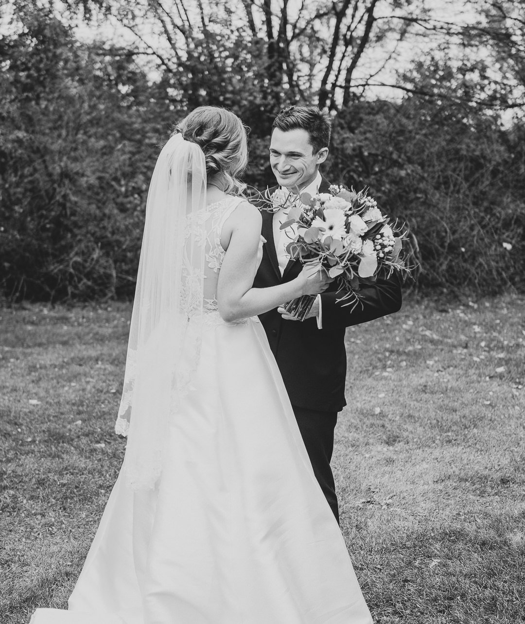 A black and white photo of a bride and groom standing outdoors, with the bride holding a large bouquet of flowers, and the groom smiling at her. The background features trees and bushes.