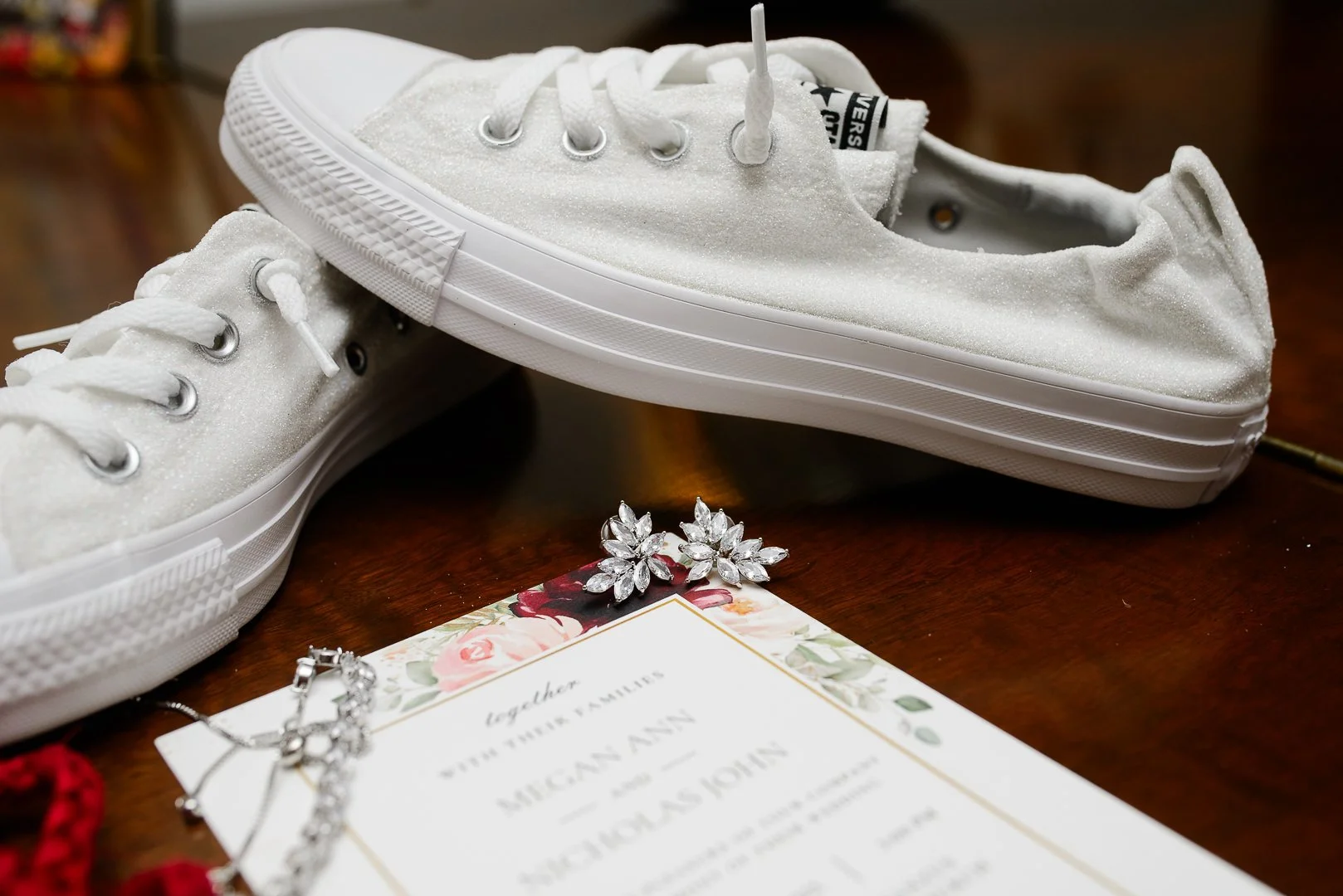 A pair of white Converse sneakers with glittery texture, jewelry including earrings and a necklace, and a wedding invitation on a wooden surface.