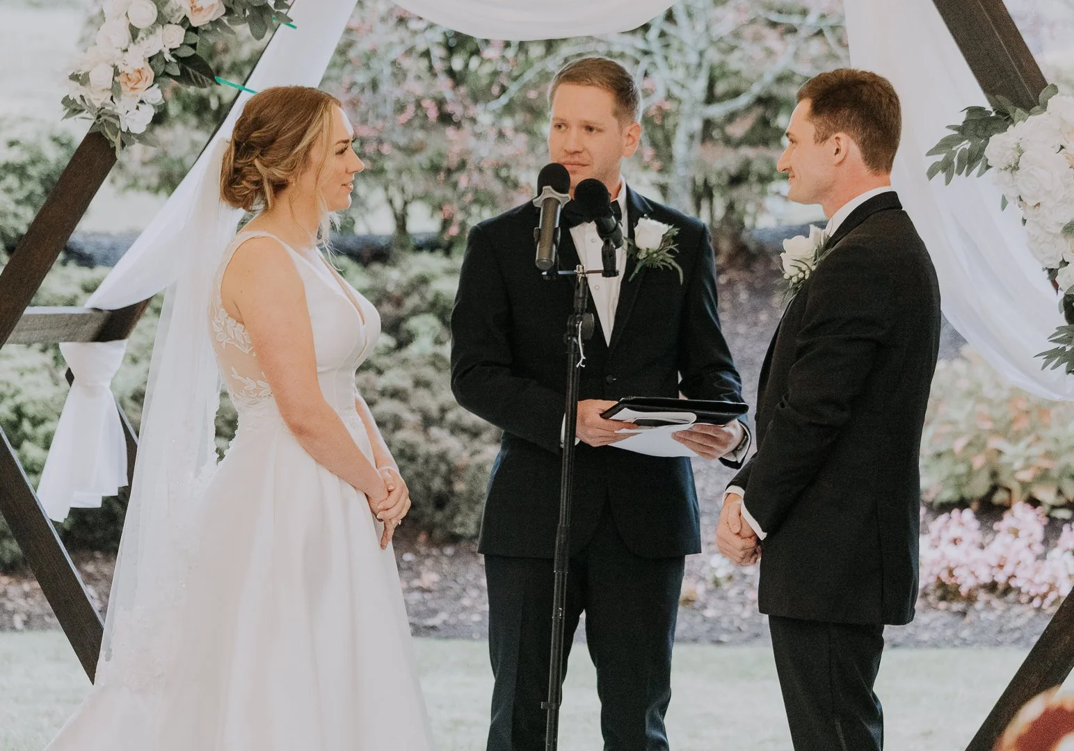 A bride and groom standing face to face during a wedding ceremony outdoors, with an officiant between them, under a decorated wooden arch with white flowers and drapery.