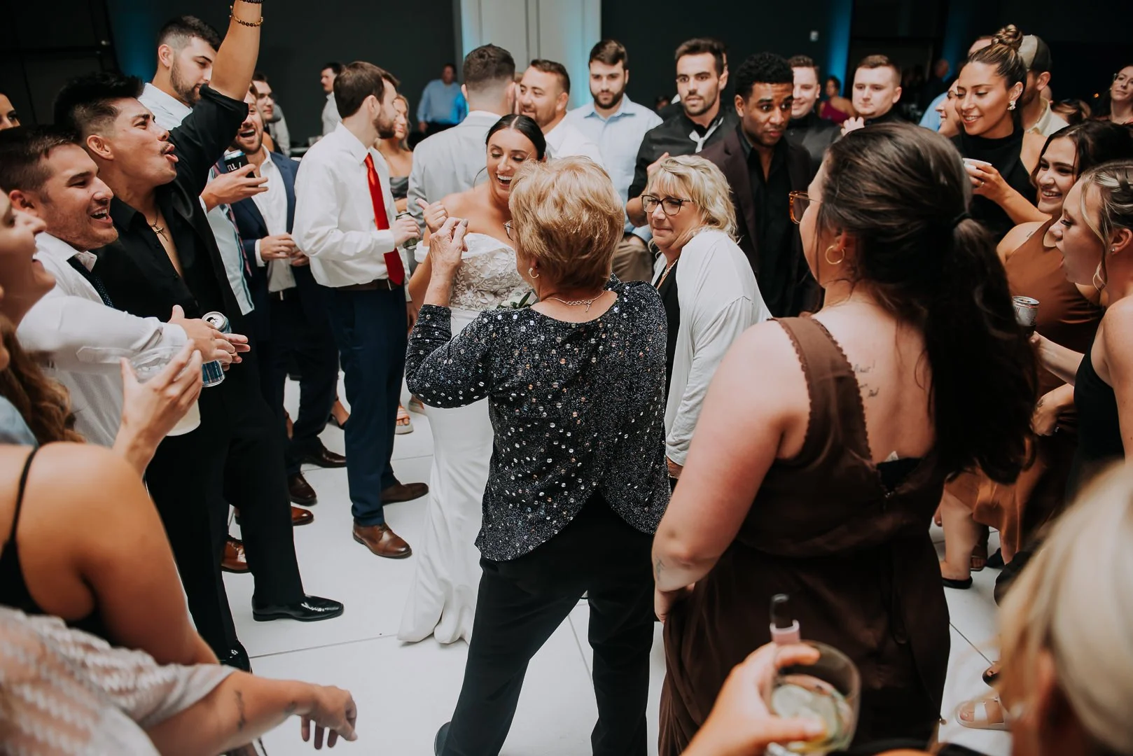 A woman in a white wedding dress smiles and dances with guests at a lively wedding reception. The crowd surrounds her, chatting and enjoying drinks.