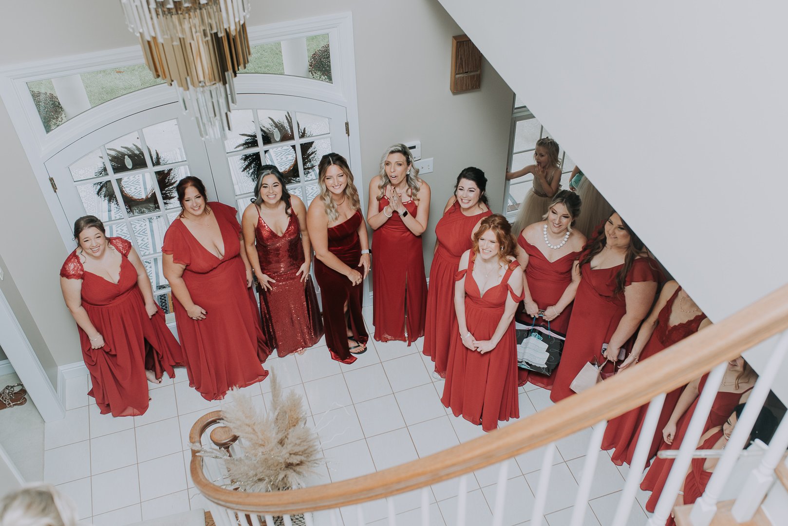 A group of women in red dresses standing in a semicircle inside a house, looking up and smiling. The view is from a staircase overlooking them.