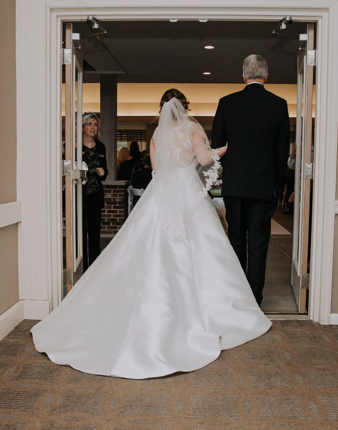 A bride in a white wedding gown and veil walking out of a building with a man, likely her father, during a wedding ceremony.