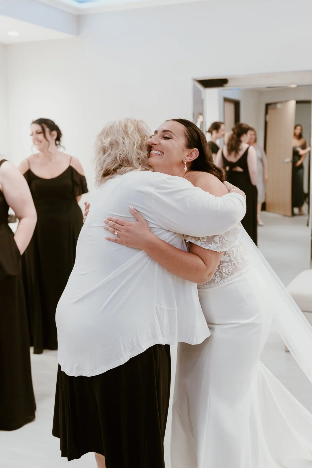 A bride in a white wedding gown hugging an older woman in a white blouse and black skirt at a wedding reception, with other women in black dresses in the background.