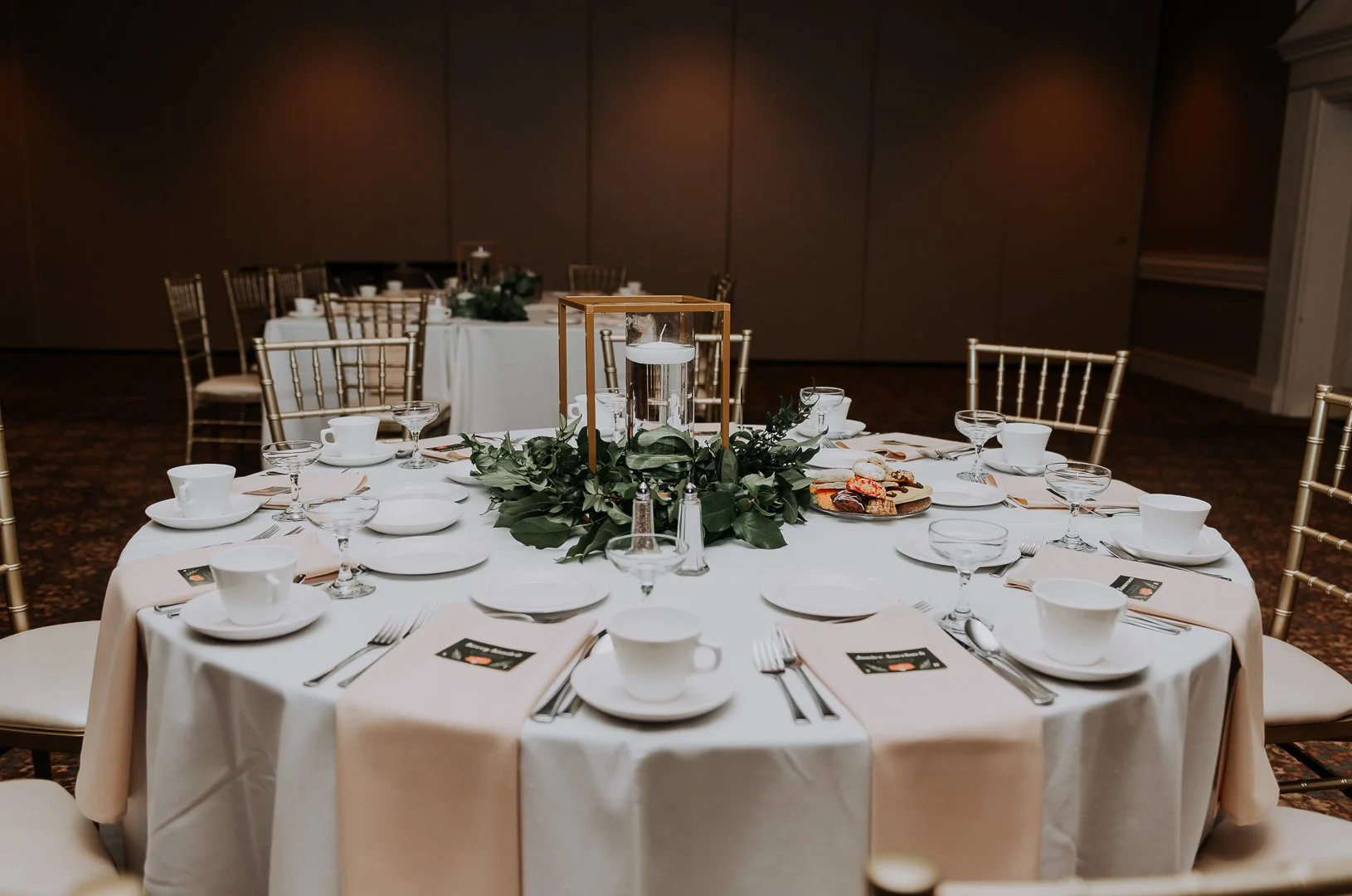 Round banquet table set with white tablecloth, white plates, cups, glasses, silverware, and a centerpiece with greenery and a water-filled glass lantern in a dimly lit event hall.