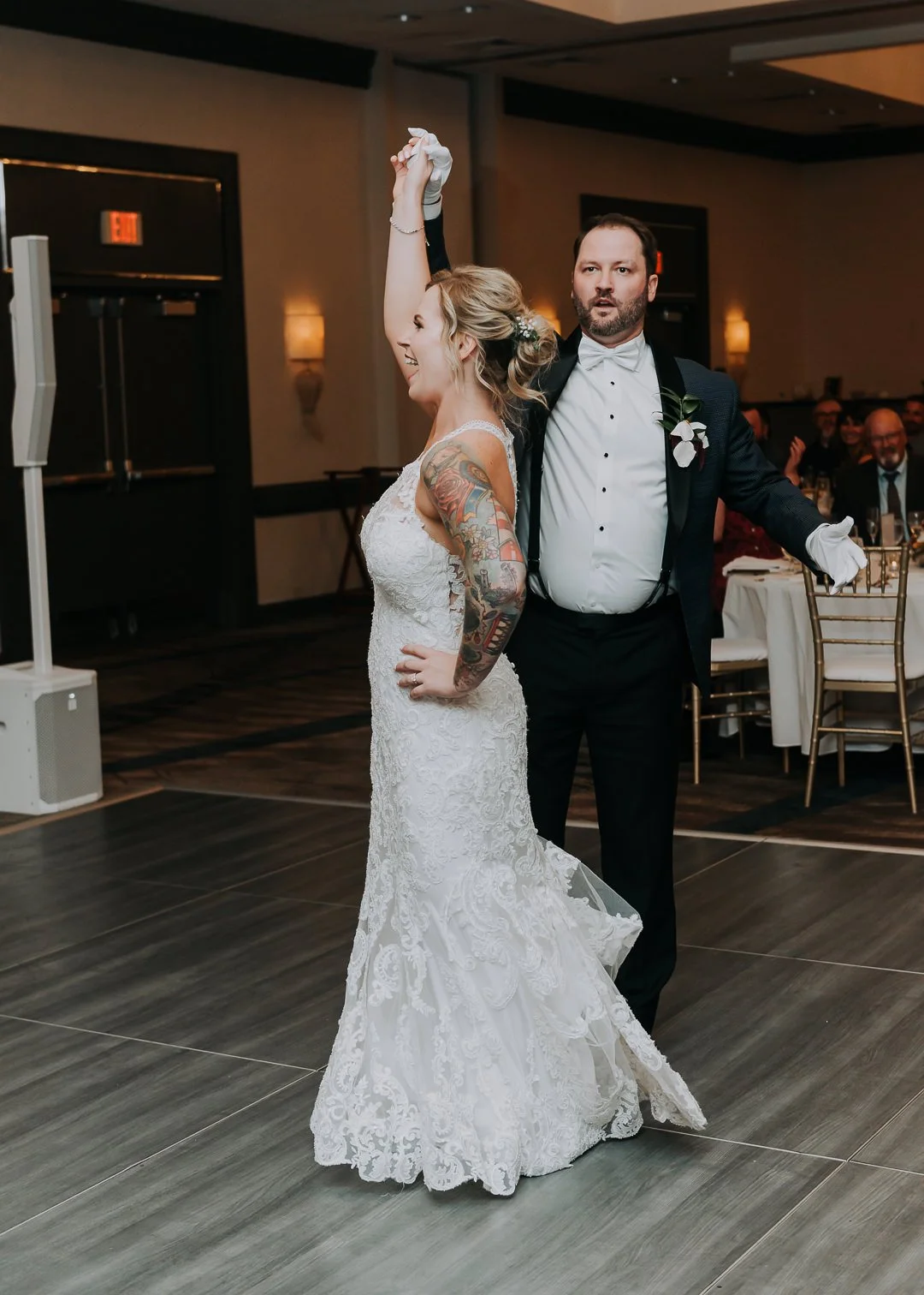 A bride with tattoos on her arm and a groom in a tuxedo dancing together at a wedding reception, with guests seated in the background.