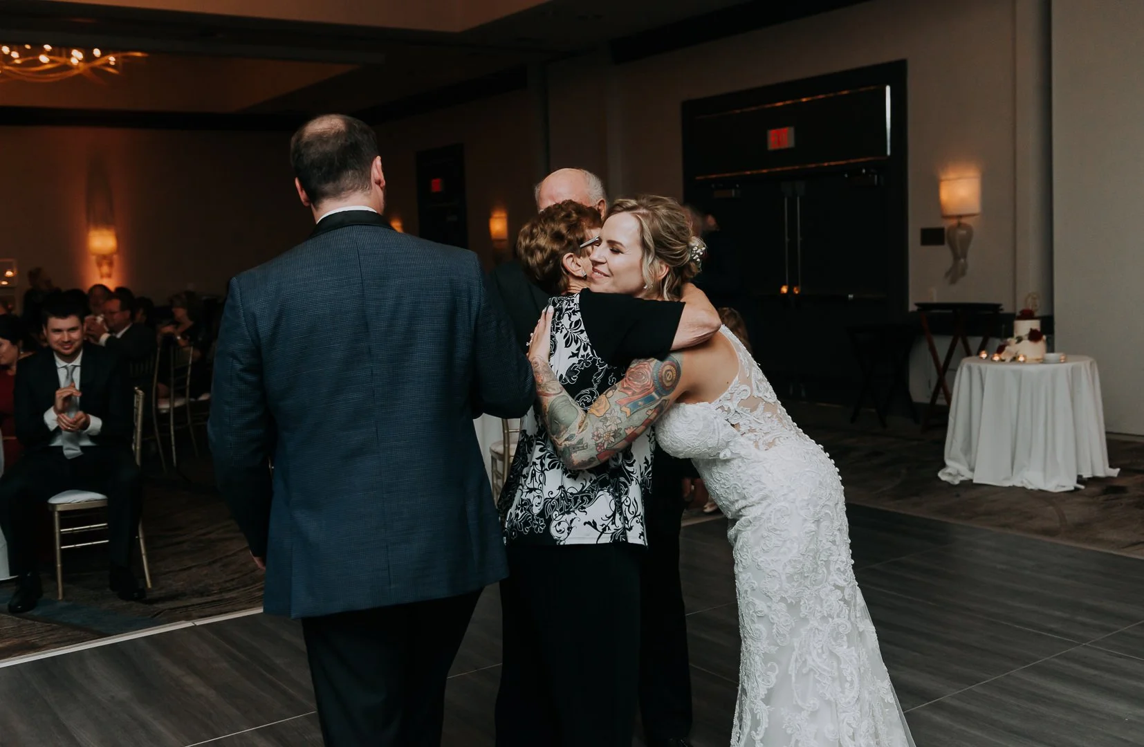 A bride hugging an older woman at her wedding reception, with a man in a suit watching, in a decorated event hall.