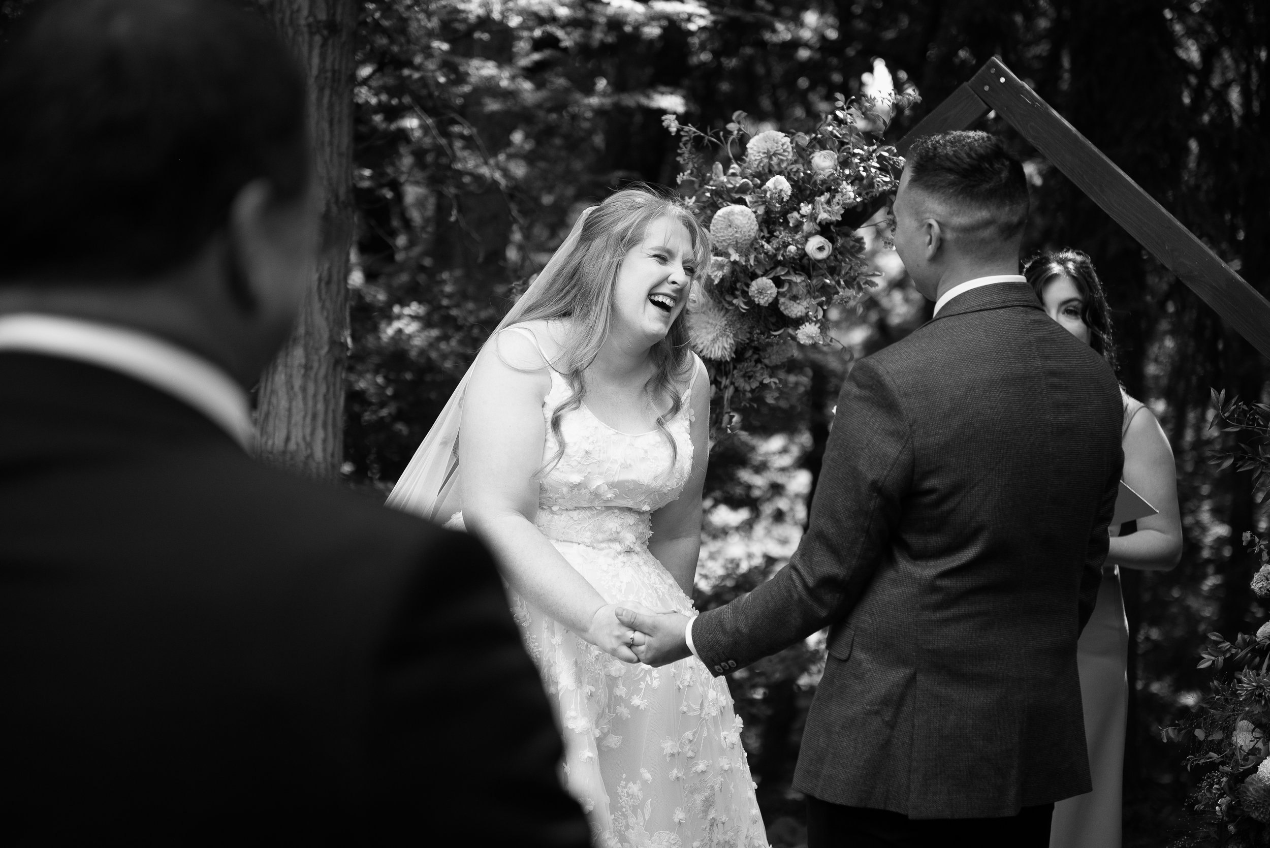 A black and white photo of a wedding ceremony outdoors with the bride and groom holding hands and exchanging vows, surrounded by friends and family, with a floral arch and trees in the background.