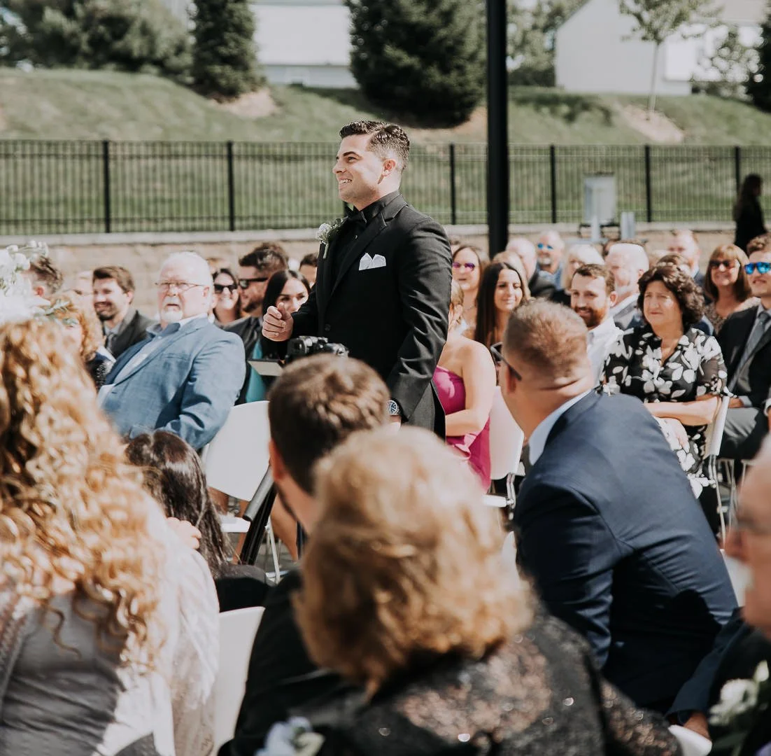 A man in a black suit standing and smiling at an outdoor wedding ceremony, surrounded by seated guests.