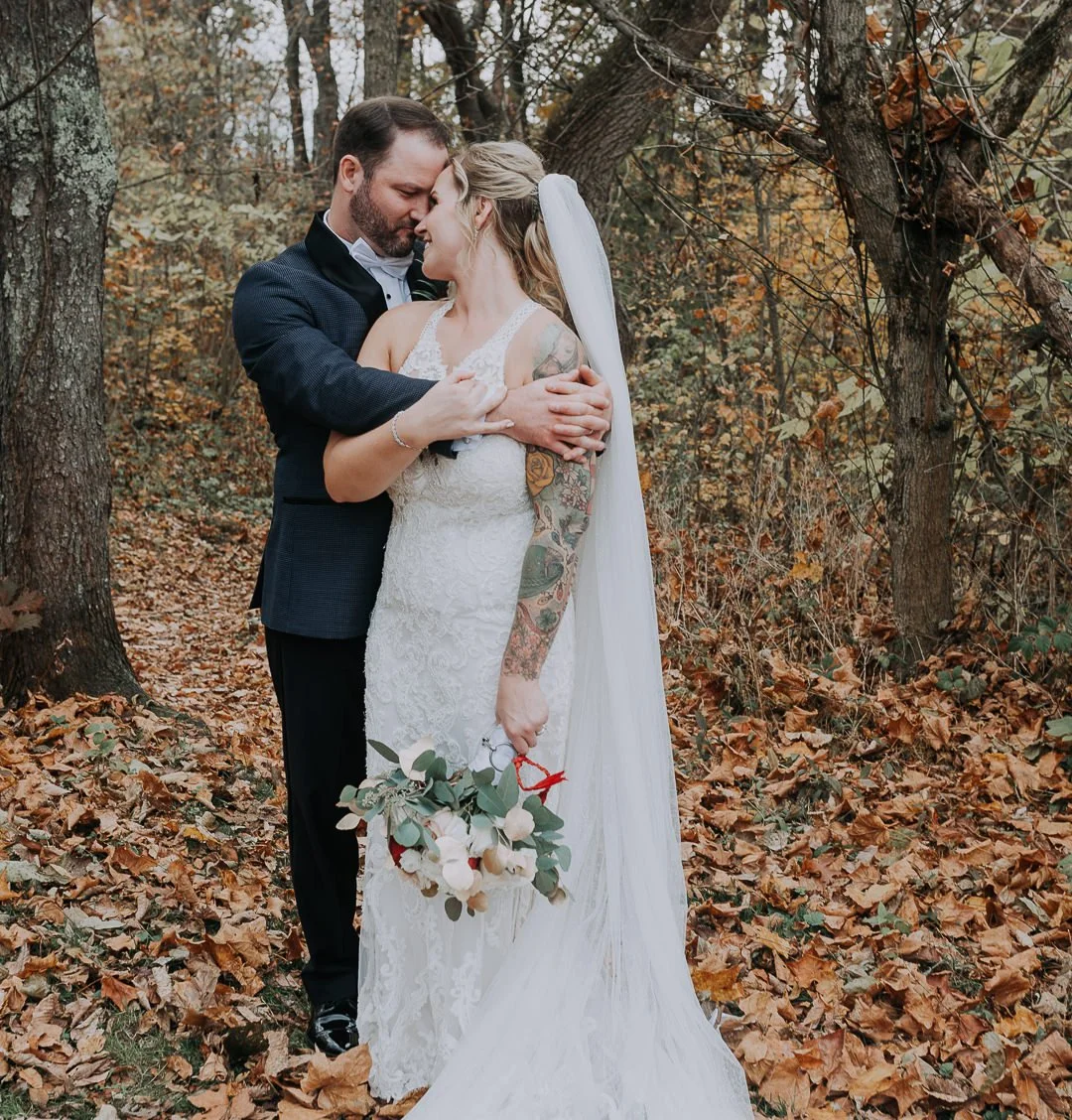 A bride and groom embracing in a forest during autumn, the bride holding a bouquet of white flowers and greenery.