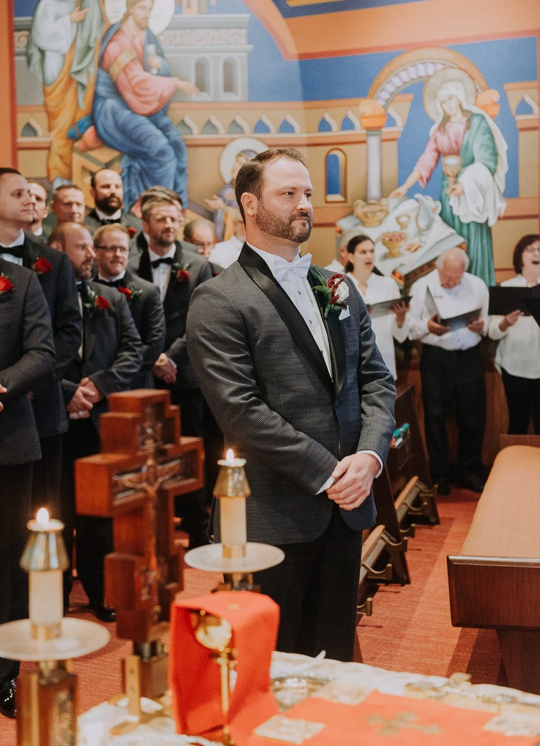 A groom with a beard and mustache, dressed in a gray suit with a white shirt and bow tie, standing inside a church during a wedding ceremony, surrounded by bridesmaids and groomsmen. The background features religious murals on the church walls.