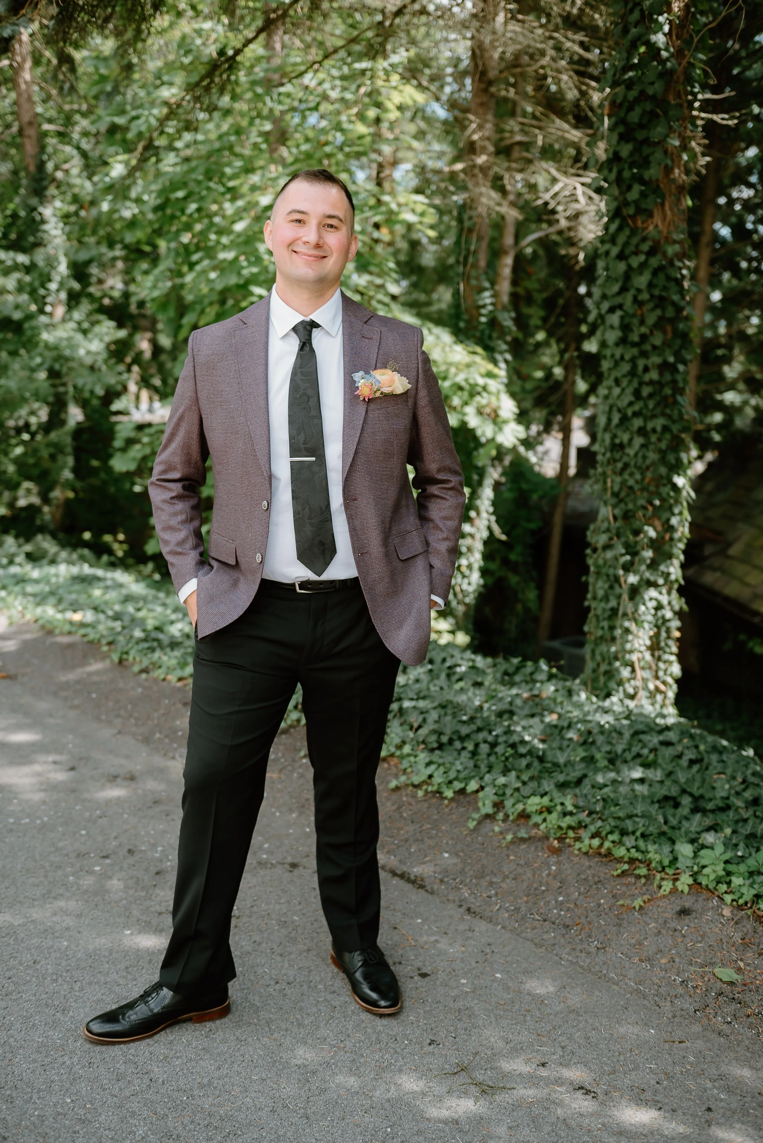 A smiling man in a suit standing outdoors among trees and ivy, with his hands in his pockets.