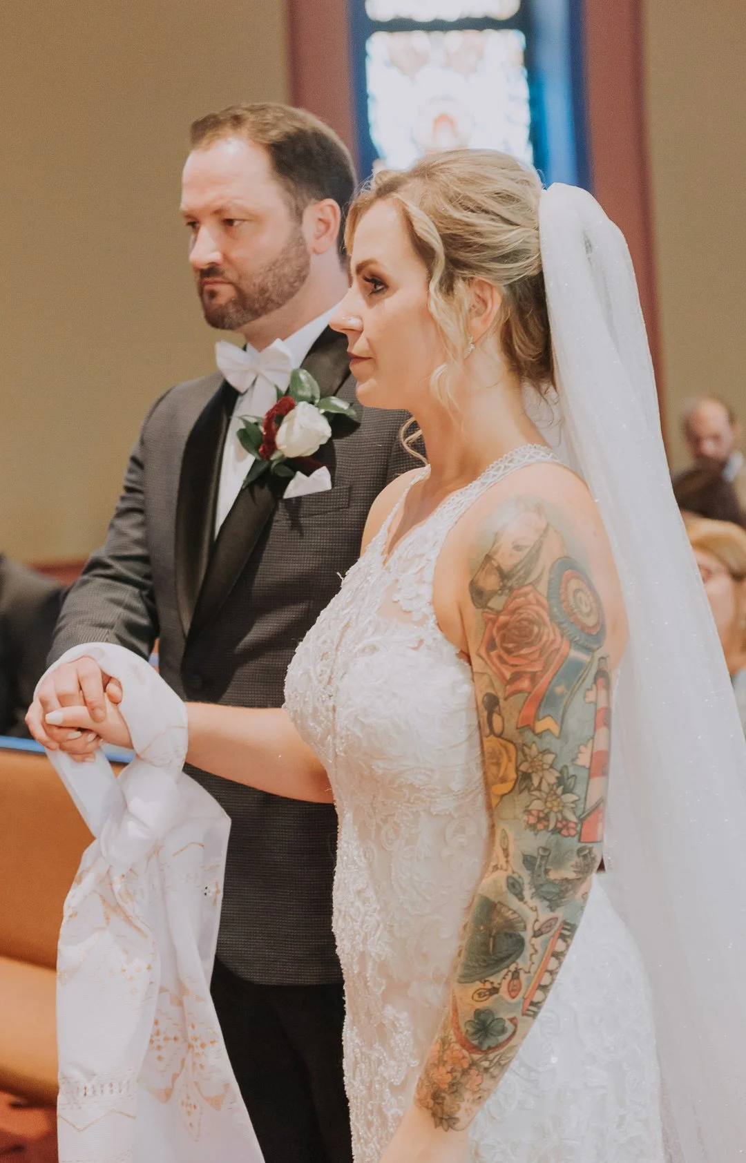 A woman with a tattooed arm wearing a white wedding dress and veil, standing next to a man in a tuxedo with a boutonniere, during a wedding ceremony in a church.