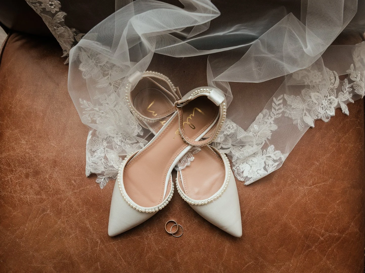 A pair of white wedding shoes with pointed toes and pearl trim, surrounded by a lace veil and two rings on a leather surface.