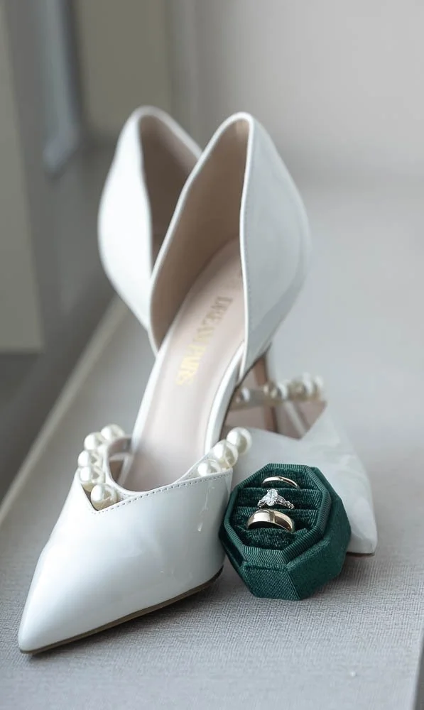 White high-heeled wedding shoes with pearl embellishments and wedding rings in a green ring box.