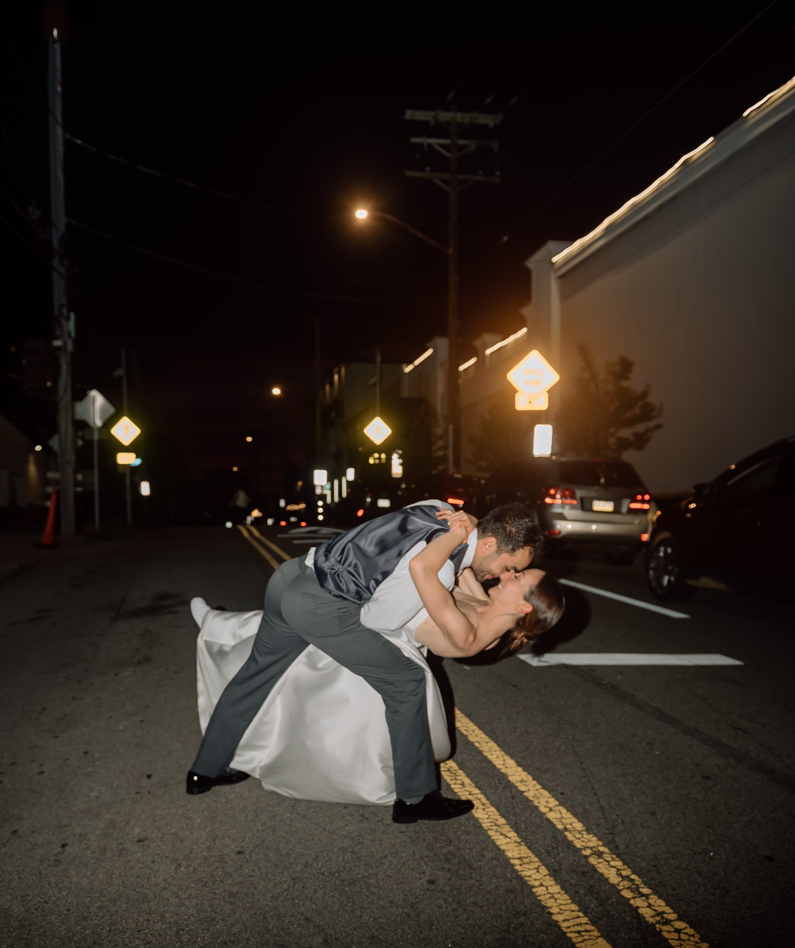 A couple dancing in the street at night, with the man dipping the woman while they hold hands and look at each other, surrounded by parked cars and streetlights.
