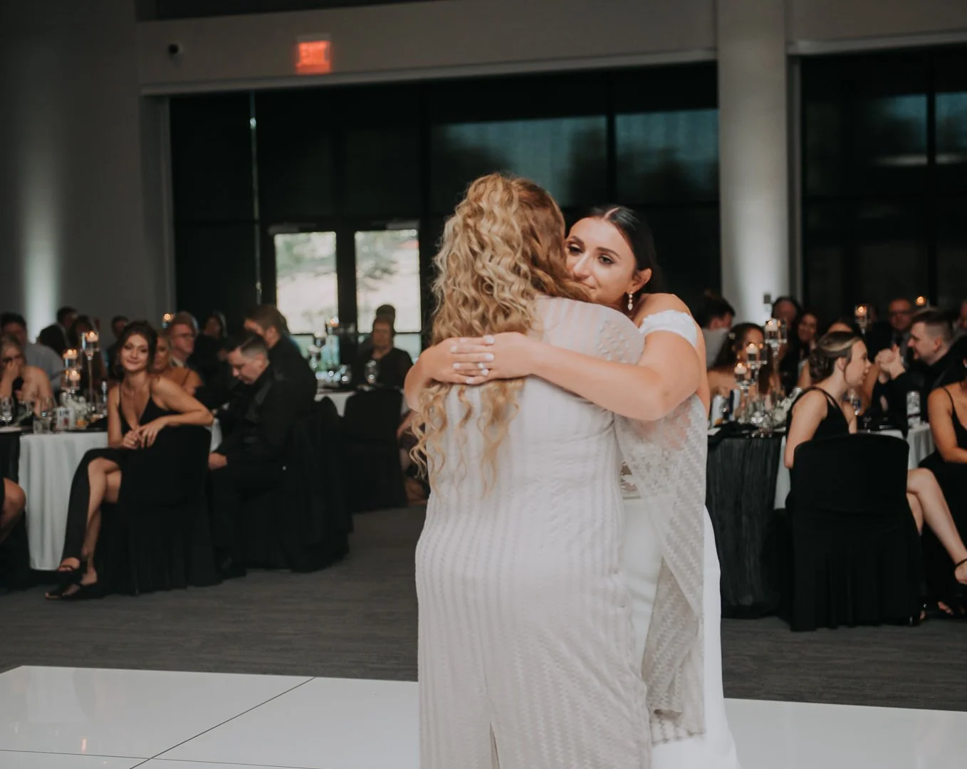 Two women wearing white dresses embracing and dancing at a wedding reception with seated guests in the background.