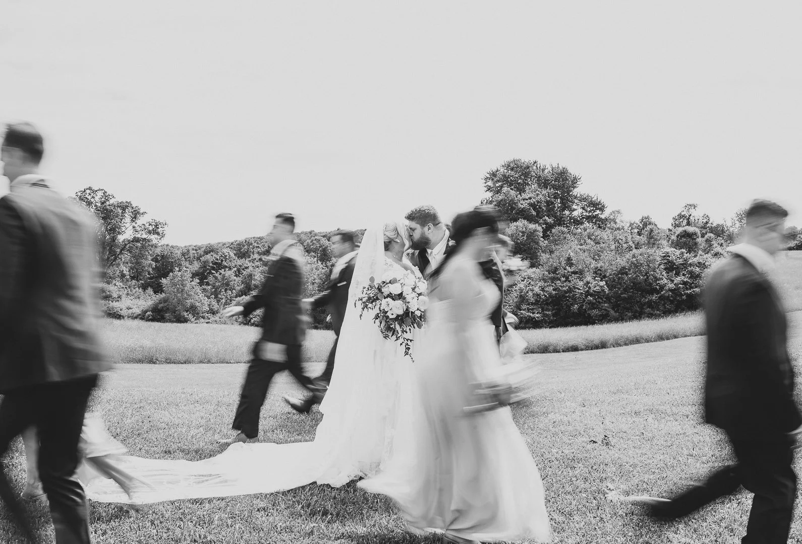 Black and white photo of a wedding ceremony outdoors with a bride and groom kissing, surrounded by people walking by, trees, and an open sky.
