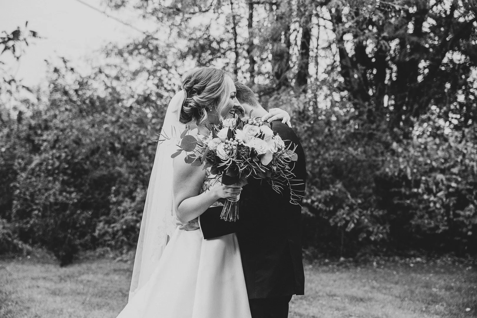 Black and white photo of a bride and groom embracing outdoors, with the bride holding a bouquet of flowers and smiling.