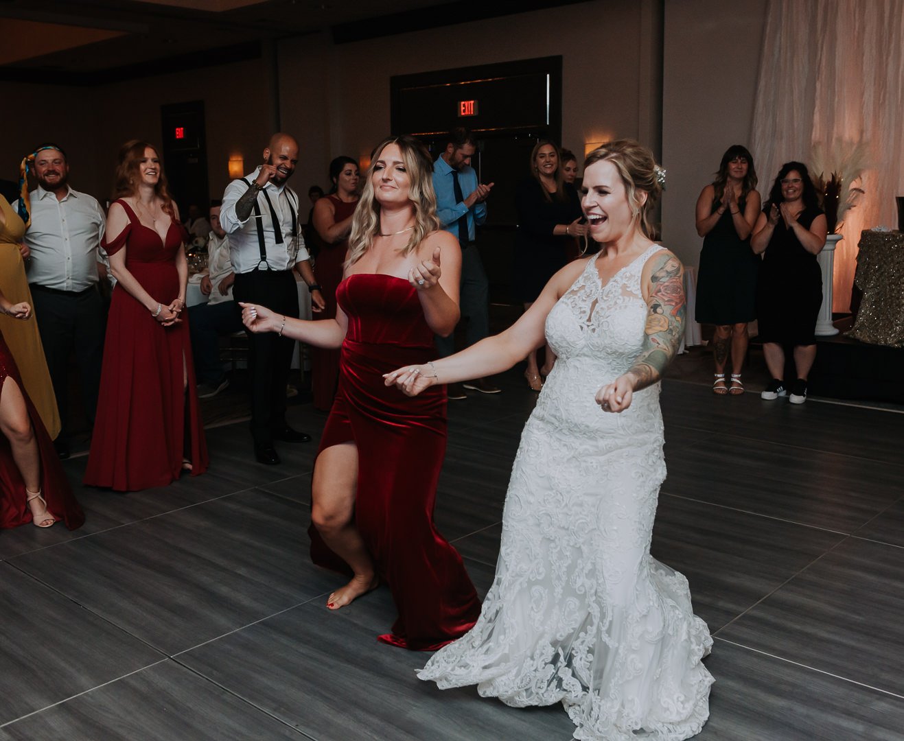 Two women, one in a white lace wedding dress and the other in a red dress, are dancing and smiling at a wedding reception, while guests look on and celebrate in the background.