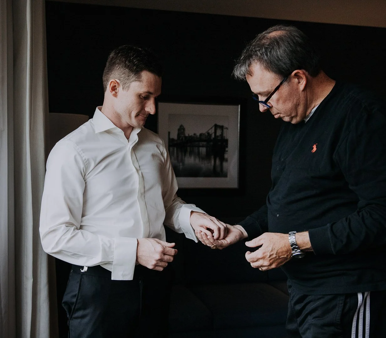 A younger man in a white shirt and black pants holding hands with an older man wearing glasses, a black shirt, and a watch, in a dimly lit room with a black and white photograph of a bridge on the wall behind them.