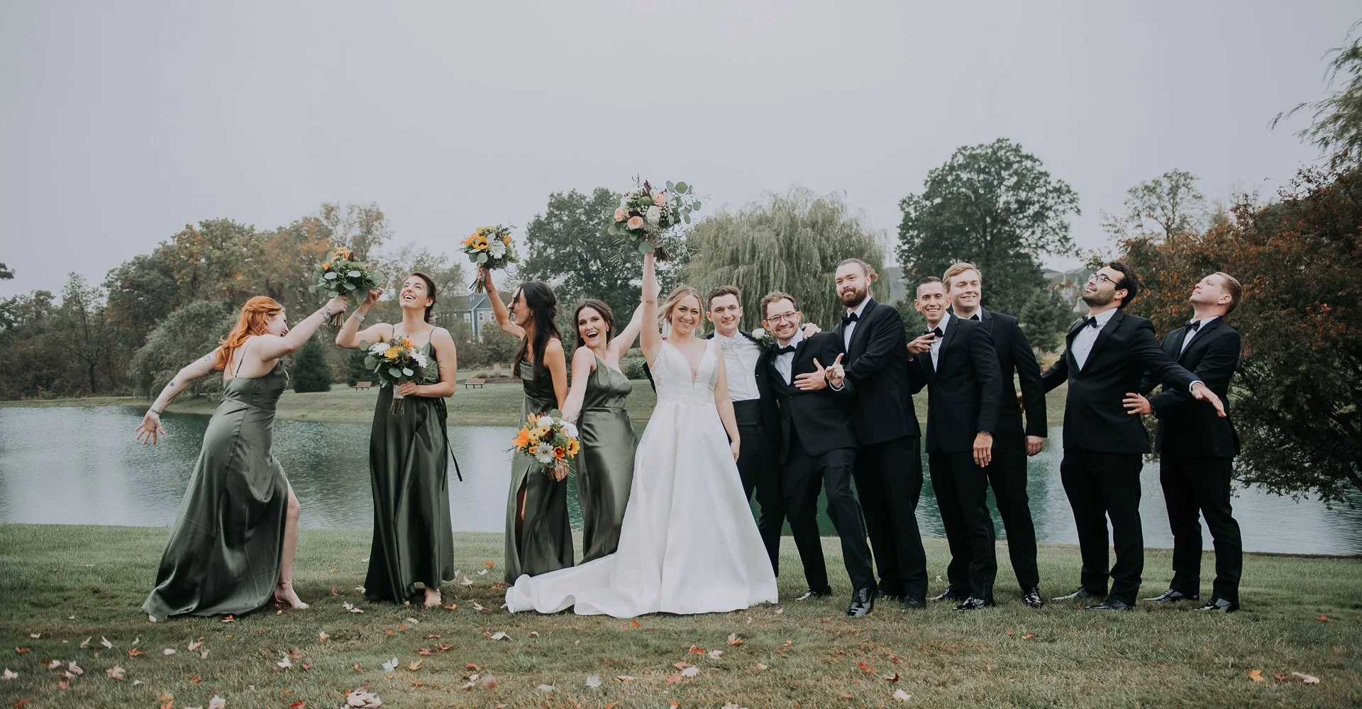Group of wedding guests, including a bride in a white gown and bridesmaids in olive green dresses, standing on grass near a lake, celebrating with bouquets and posing joyfully on a cloudy day.