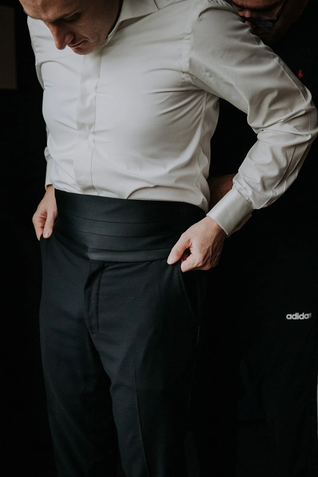 Two men, one wearing a white shirt and black pants, are helping the other put on a belt. The man in the white shirt has his hands on his hips and is looking down. The other man is partially visible behind him, also assisting.